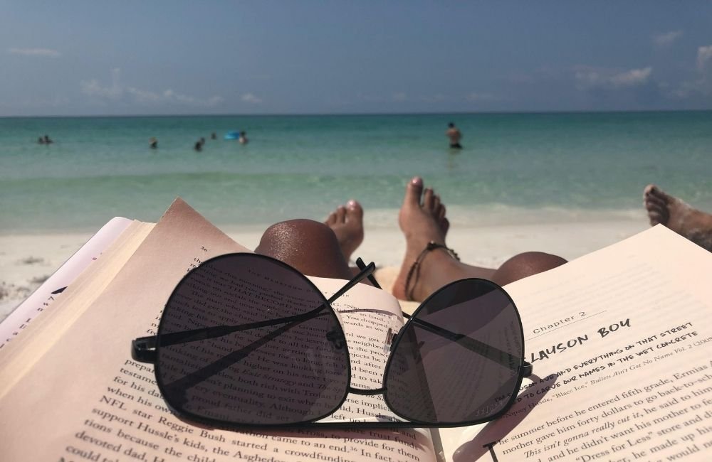 A person laying on the beach with their book and sunglasses on the book. Give yourself permission to rest without guilt and silence your inner critic.
