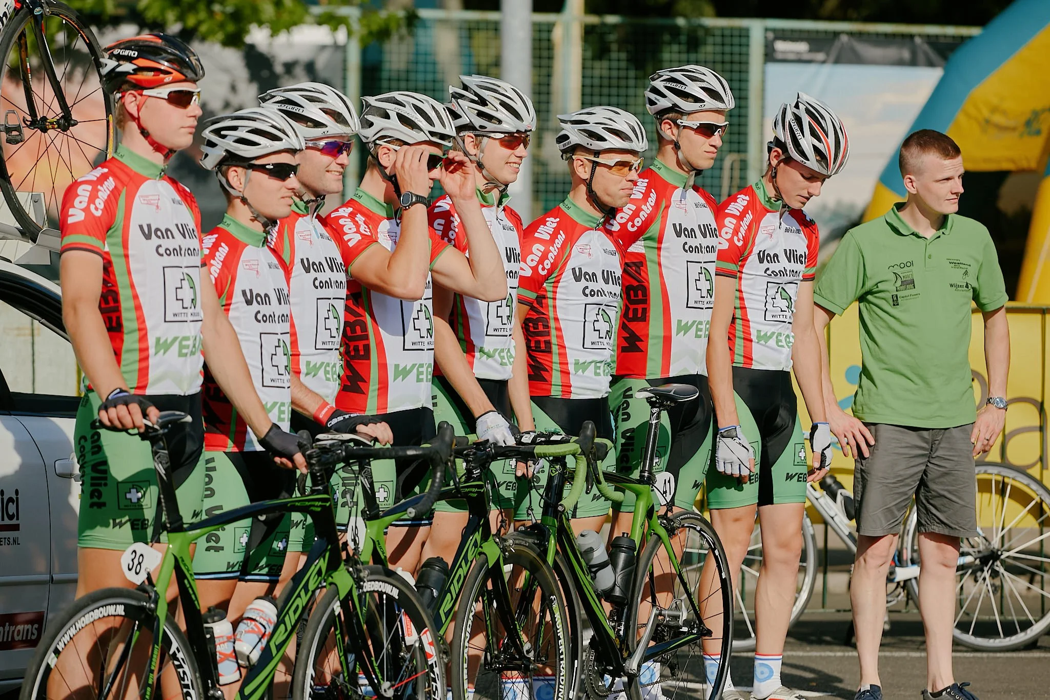 A group of cyclists wearing matching team jerseys and helmets standing at the starting line of a race with their bikes. Some are adjusting their gear, others are looking ahead, in an outdoor setting.