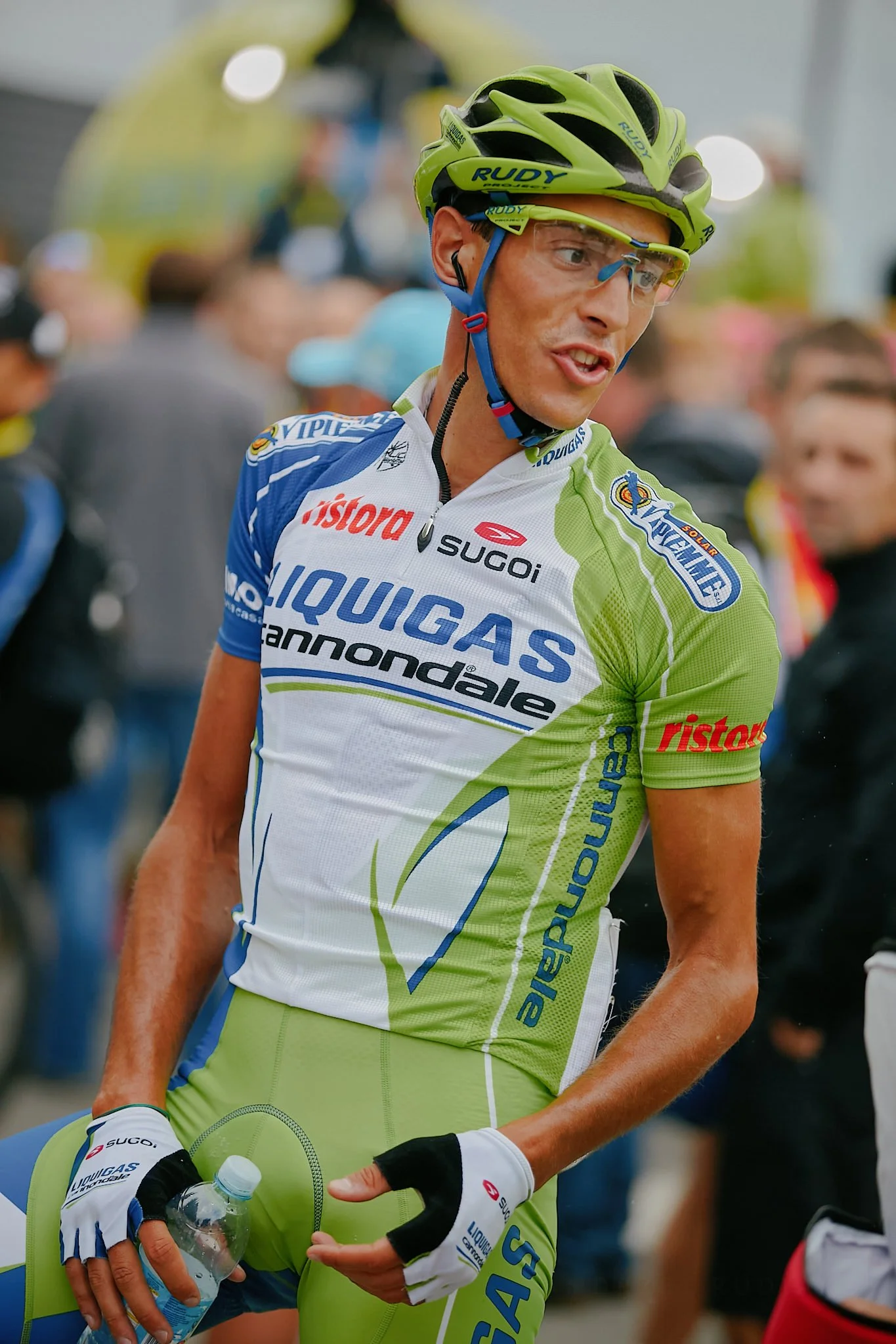 A male cyclist in green and white race gear, wearing a green helmet and protective glasses, holding a water bottle, with a blurred crowd in the background.