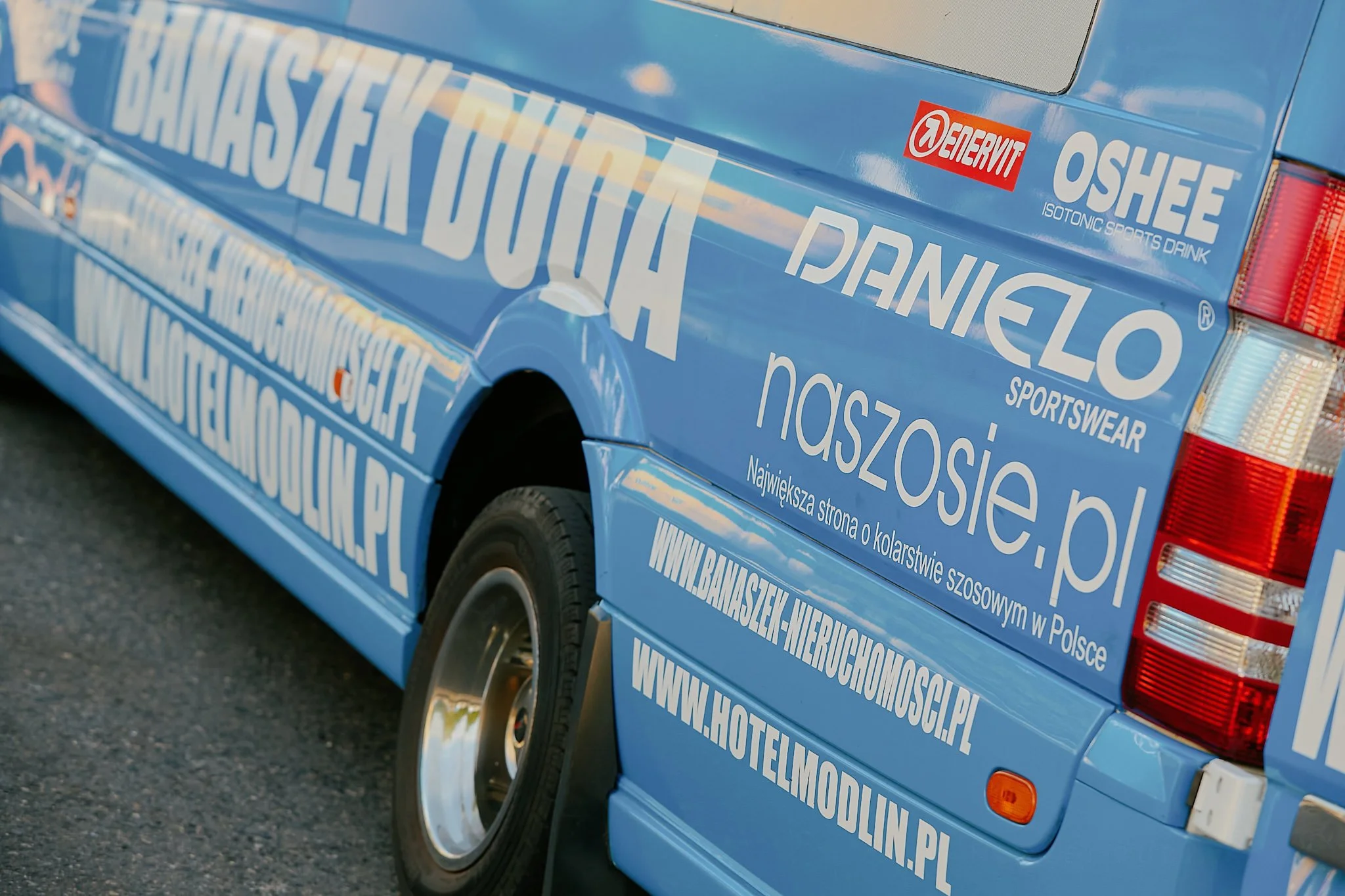 A blue vehicle with numerous white and red stickers and lettering promoting a real estate website in Polish. The vehicle features a large website URL and various brand logos, including Demerit and Oshee.
