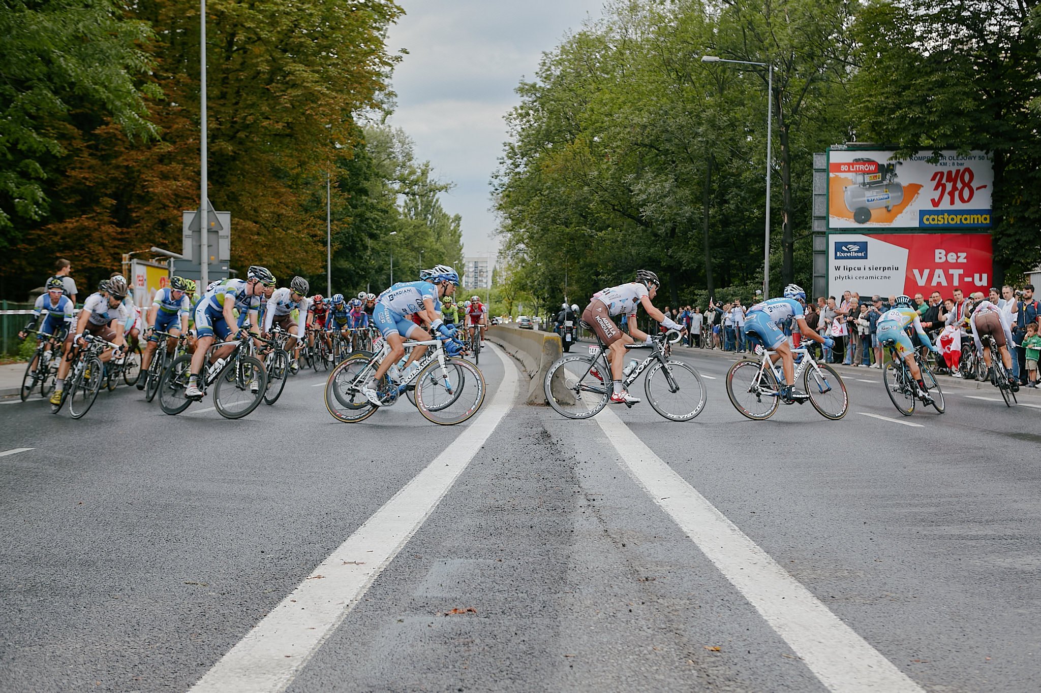 Cyclists in a race crossing a street with spectators watching on the side, surrounded by trees and advertisements.