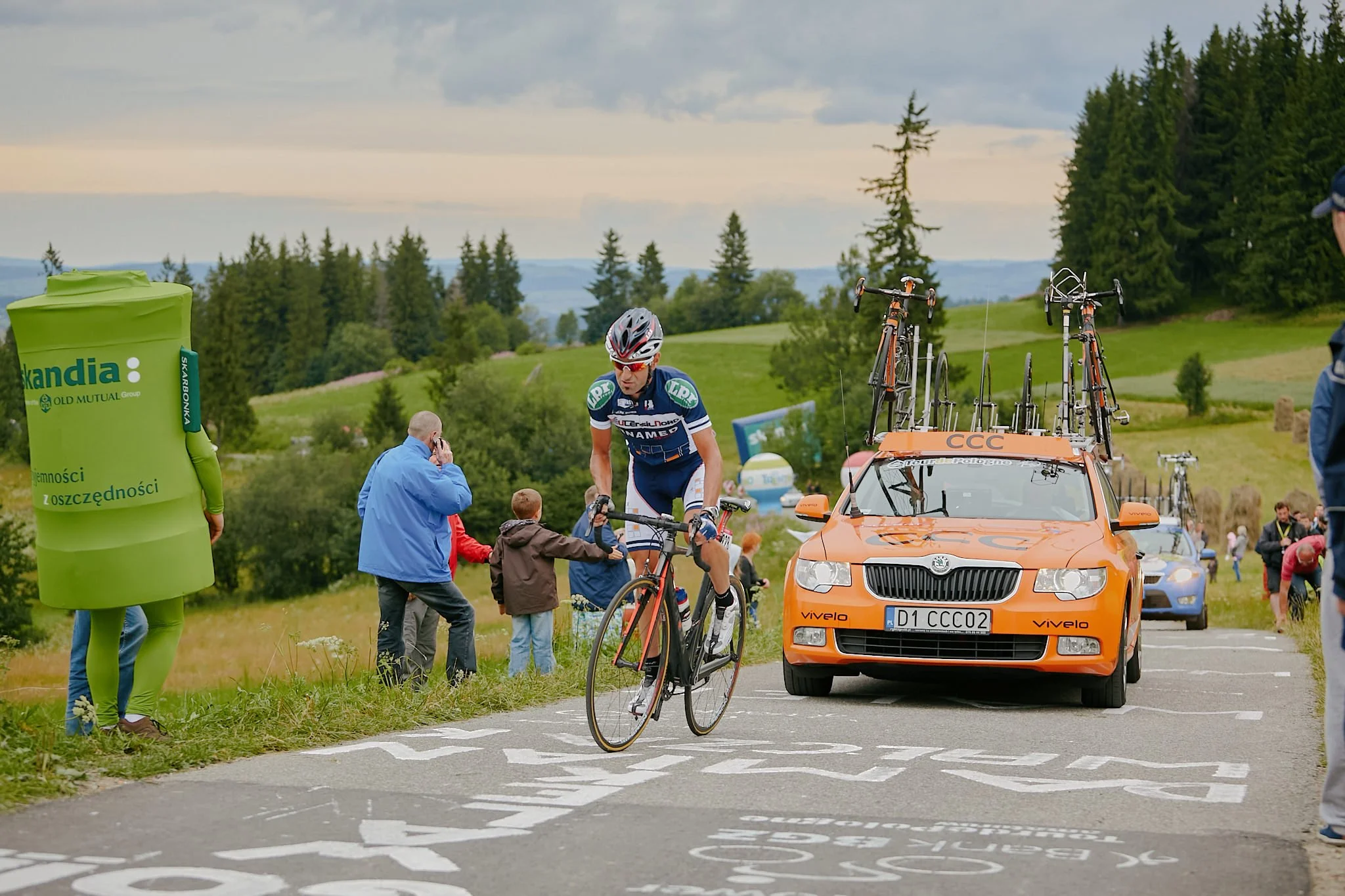 A cyclist wearing a blue and white jersey and a helmet crossing a finish line marked with white lines and text on the road, with a support car following behind on a rural road, and spectators and officials nearby, with green trees and rolling hills i