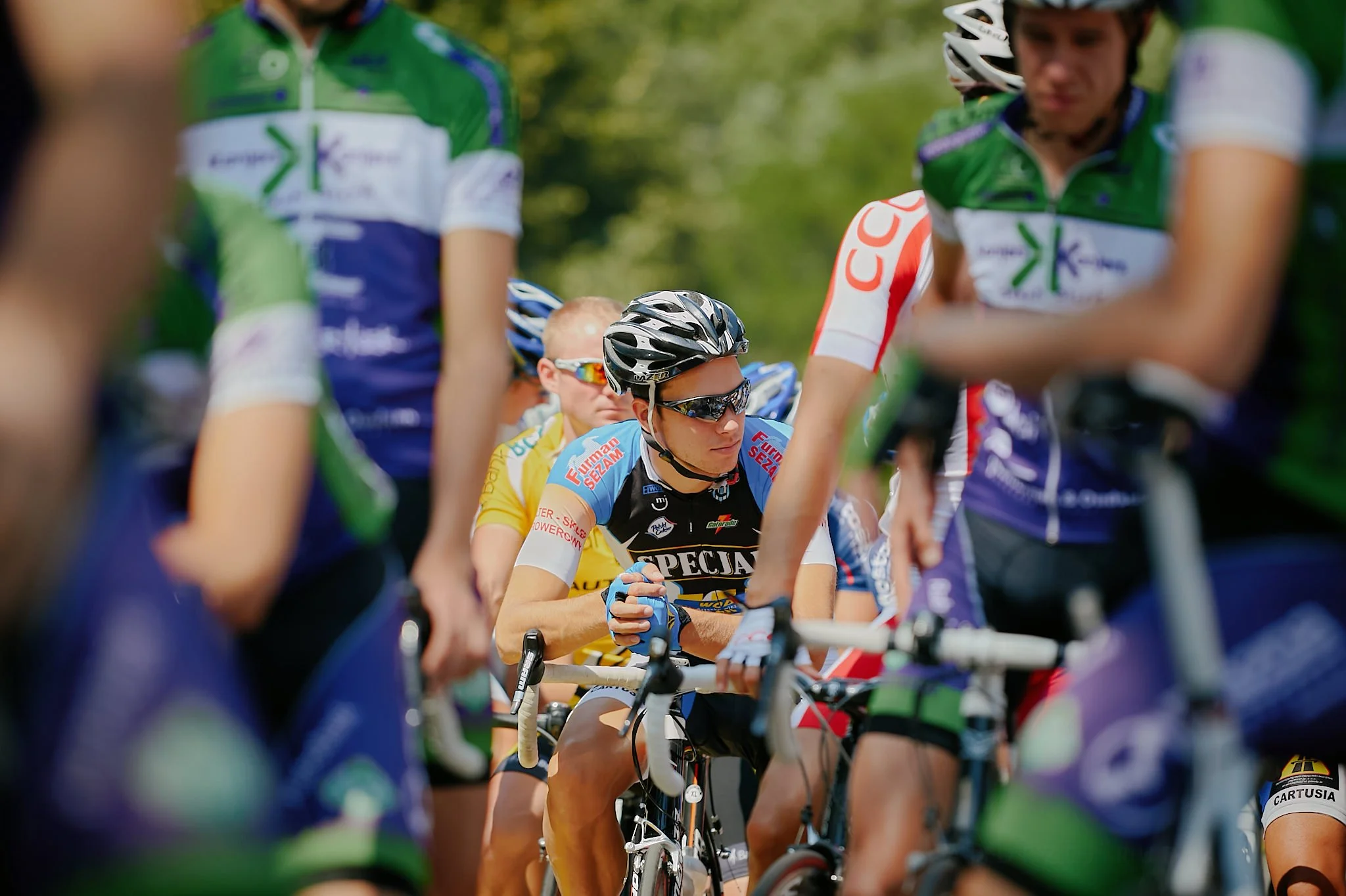 Cyclists gathered at a race event, wearing colorful uniforms and helmets, with some preparing for or participating in a cycling competition outdoors.
