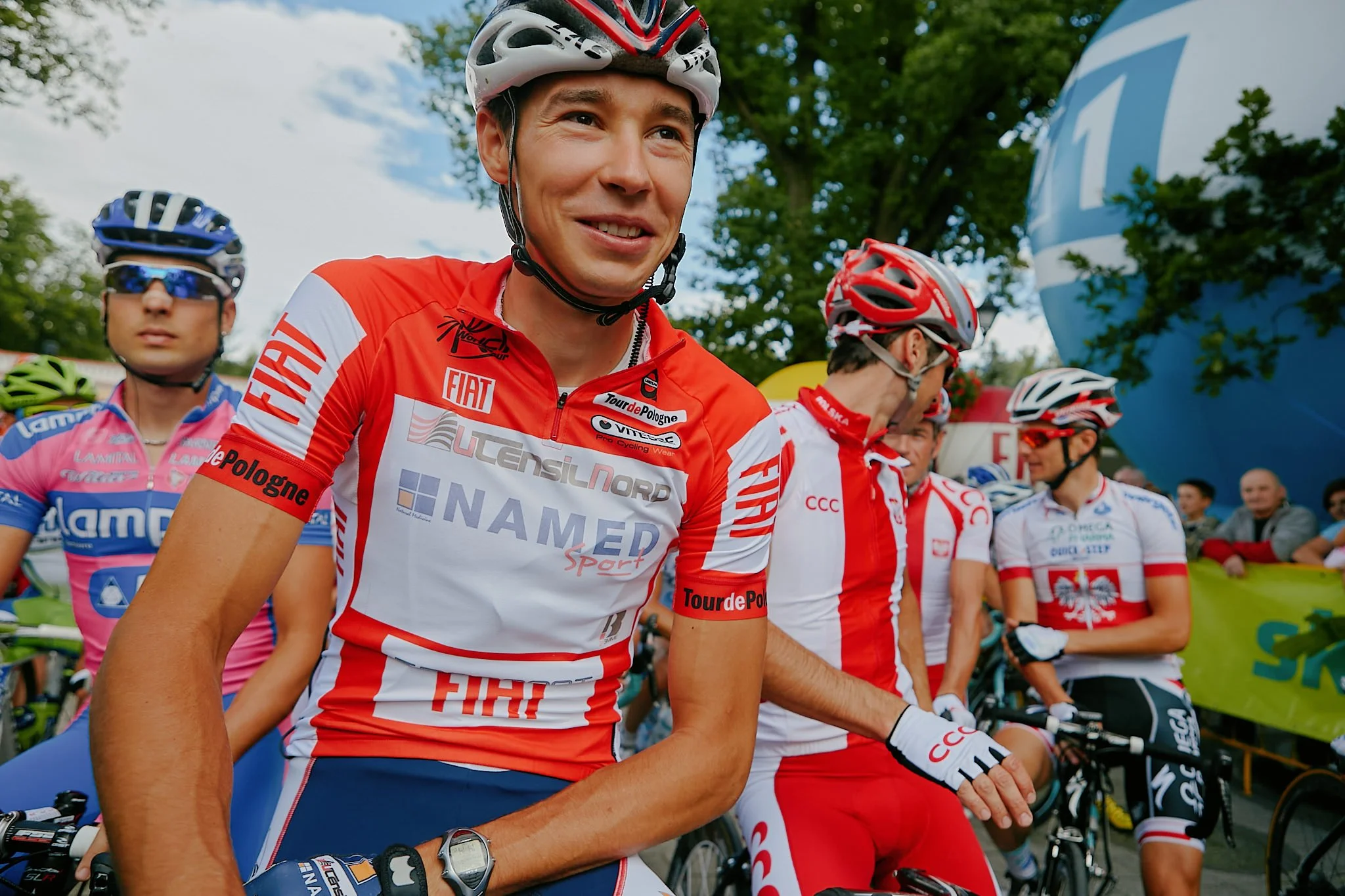 Group of cyclists in team uniforms at a cycling event, with some wearing helmets and sunglasses and one smiling person in the foreground. Trees and banners are in the background.