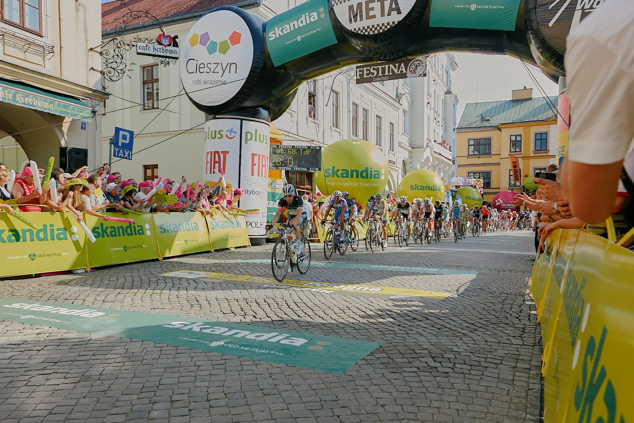 Cyclists in a race crossing the finish line in a city square, with crowds of spectators and large yellow banners with green and blue logos, and decorative advertising banners overhead.