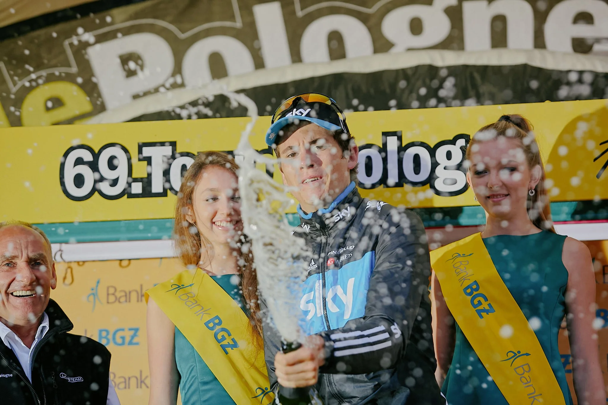 Cyclist in black and blue jersey on a podium celebrating with champagne, surrounded by two women in yellow sashes and an older man in black jacket, with a yellow background banner and water spray in the air.