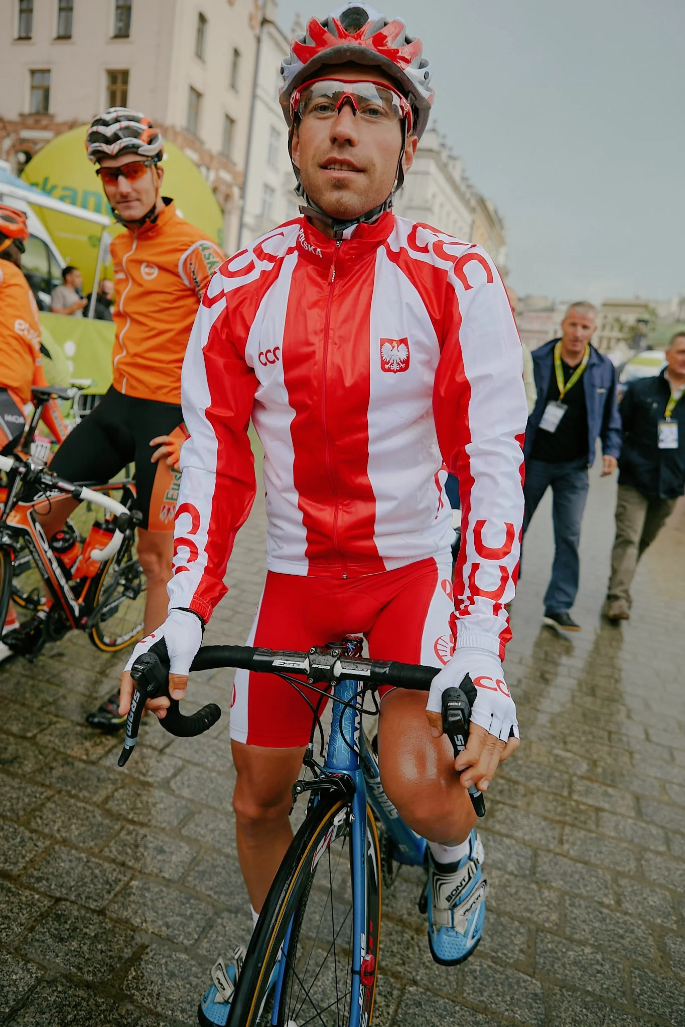 A male cyclist wearing a red and white Poland national team cycling uniform, red helmet, and sunglasses, holding a blue road bike, with people and buildings in the background.
