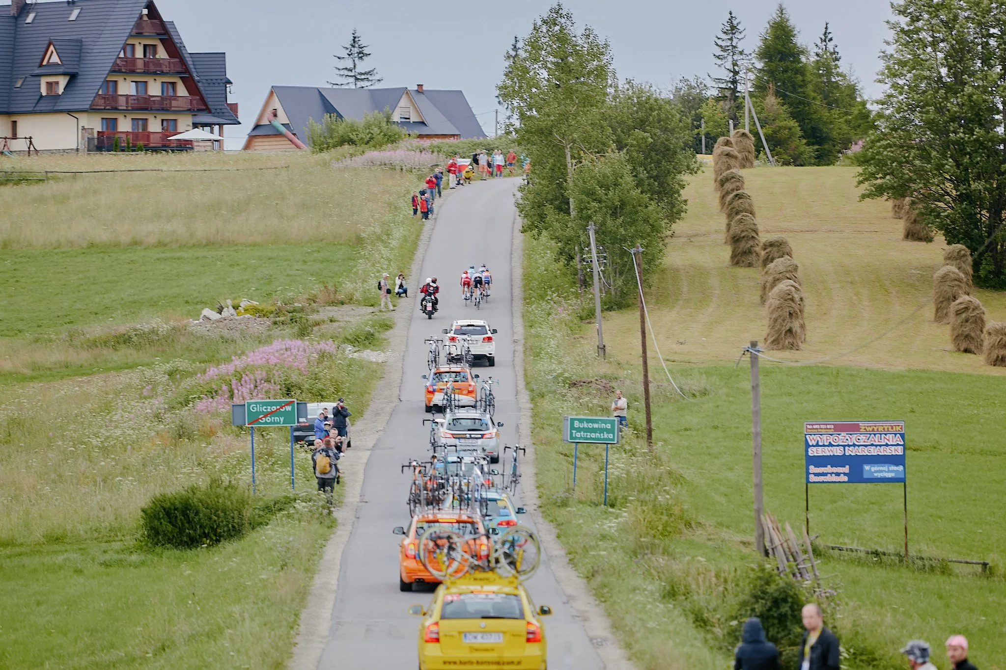 A rural scene with a long line of vehicles, including cars, trucks, and bicycles, traveling on a narrow paved road through green fields. There are groups of people walking and standing along the roadside, some with bicycles. The background features h