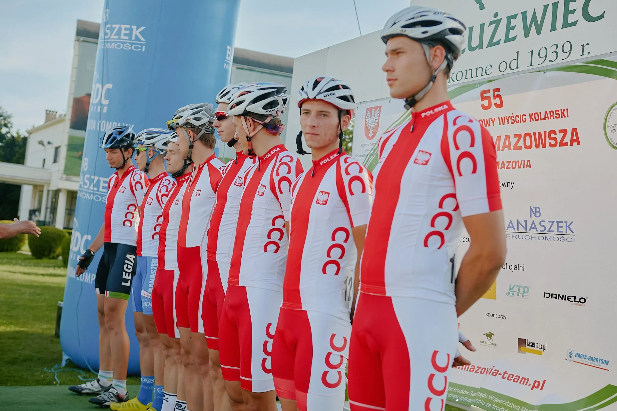 A group of young male cyclists in cycling uniforms standing in a line in front of a banner, possibly at a race event. They are wearing helmets and sunglasses, with some looking serious or focused.