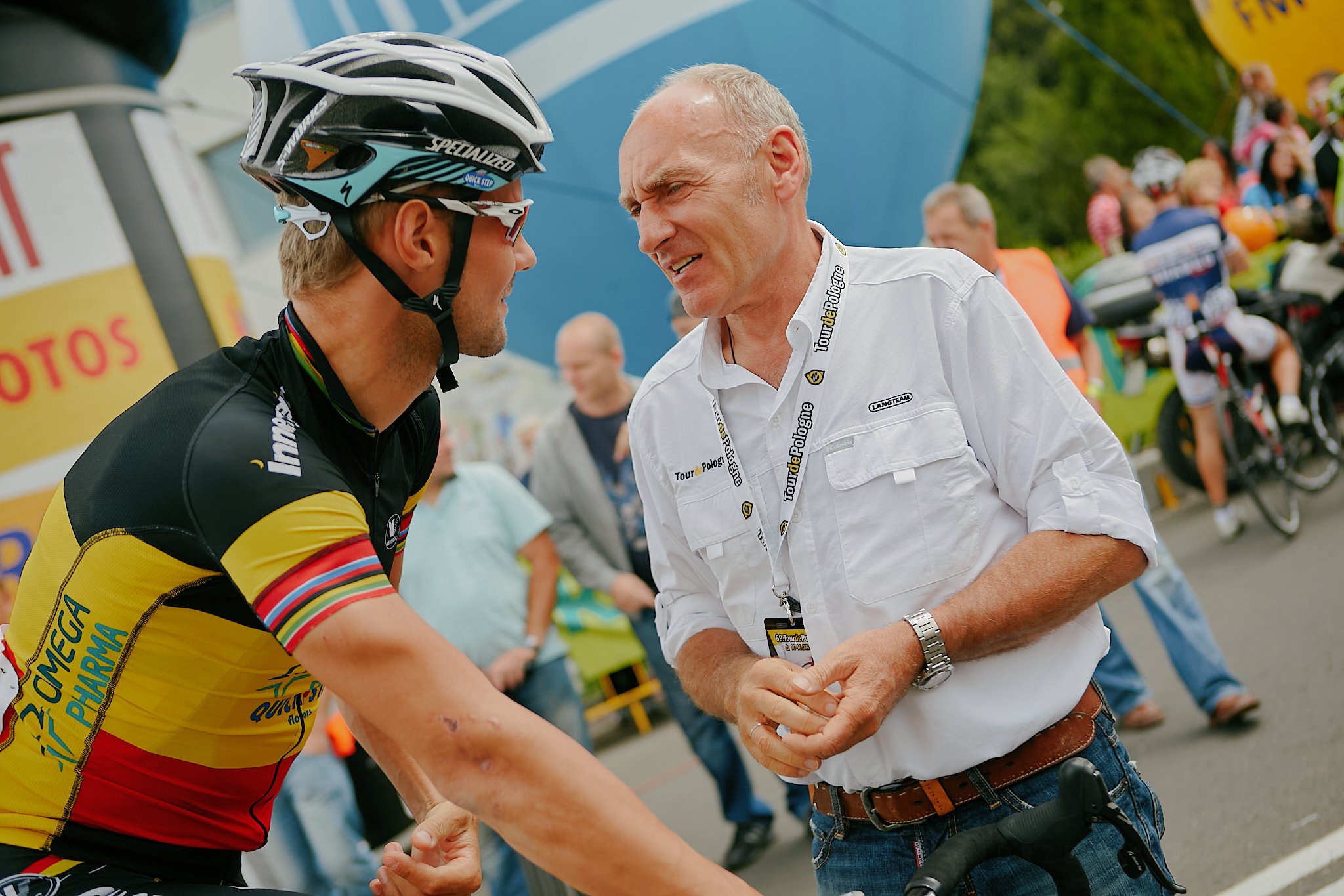 A cyclist in a yellow and black jersey shaking hands and talking with a man in a white shirt at an outdoor event, with other people and bicycles in the background.