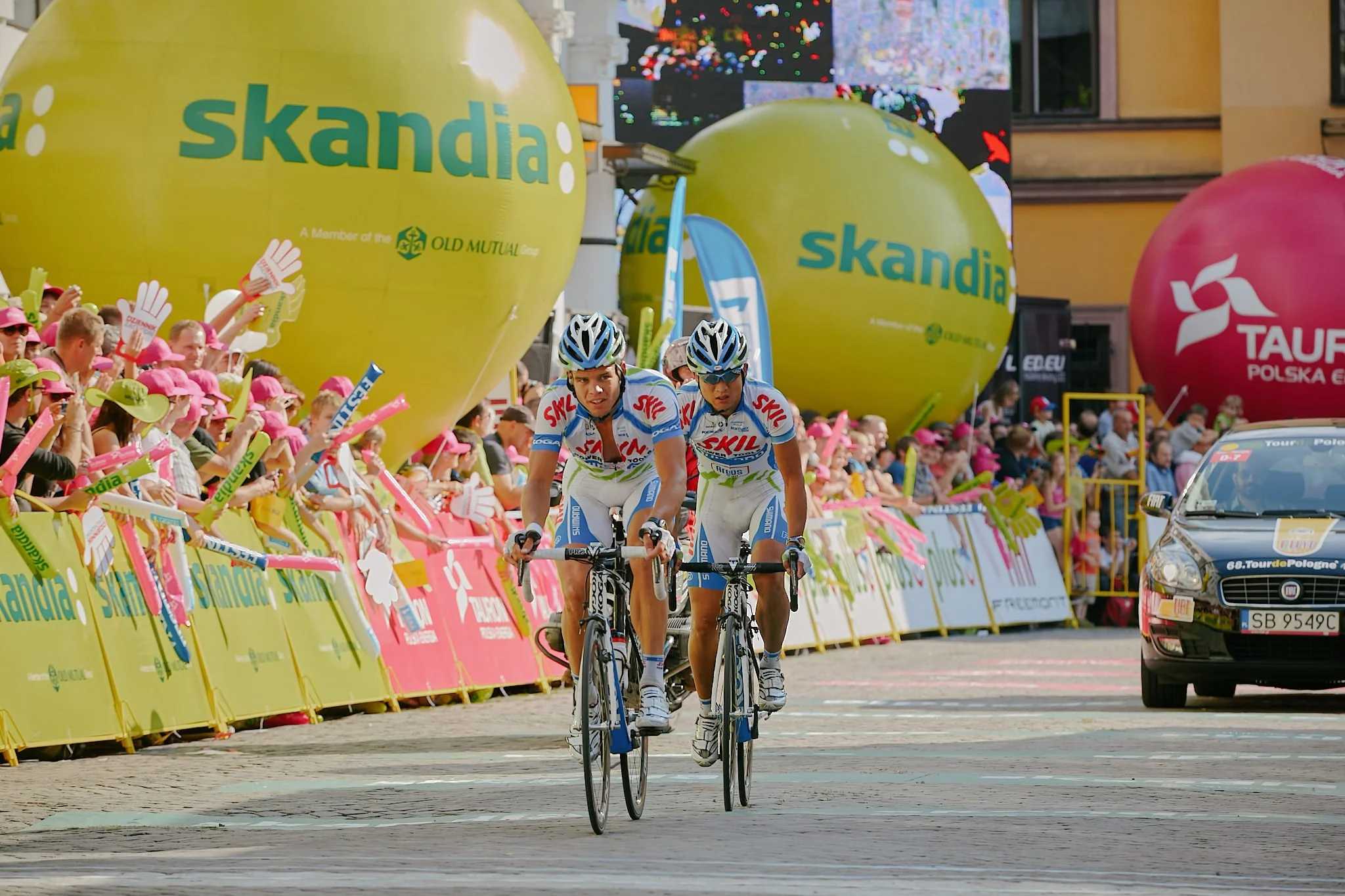 Two male cyclists in white and blue team jerseys riding bikes on a city street during a race, with a crowd of spectators behind a yellow barrier, large yellow balloons, and colorful banners in the background.