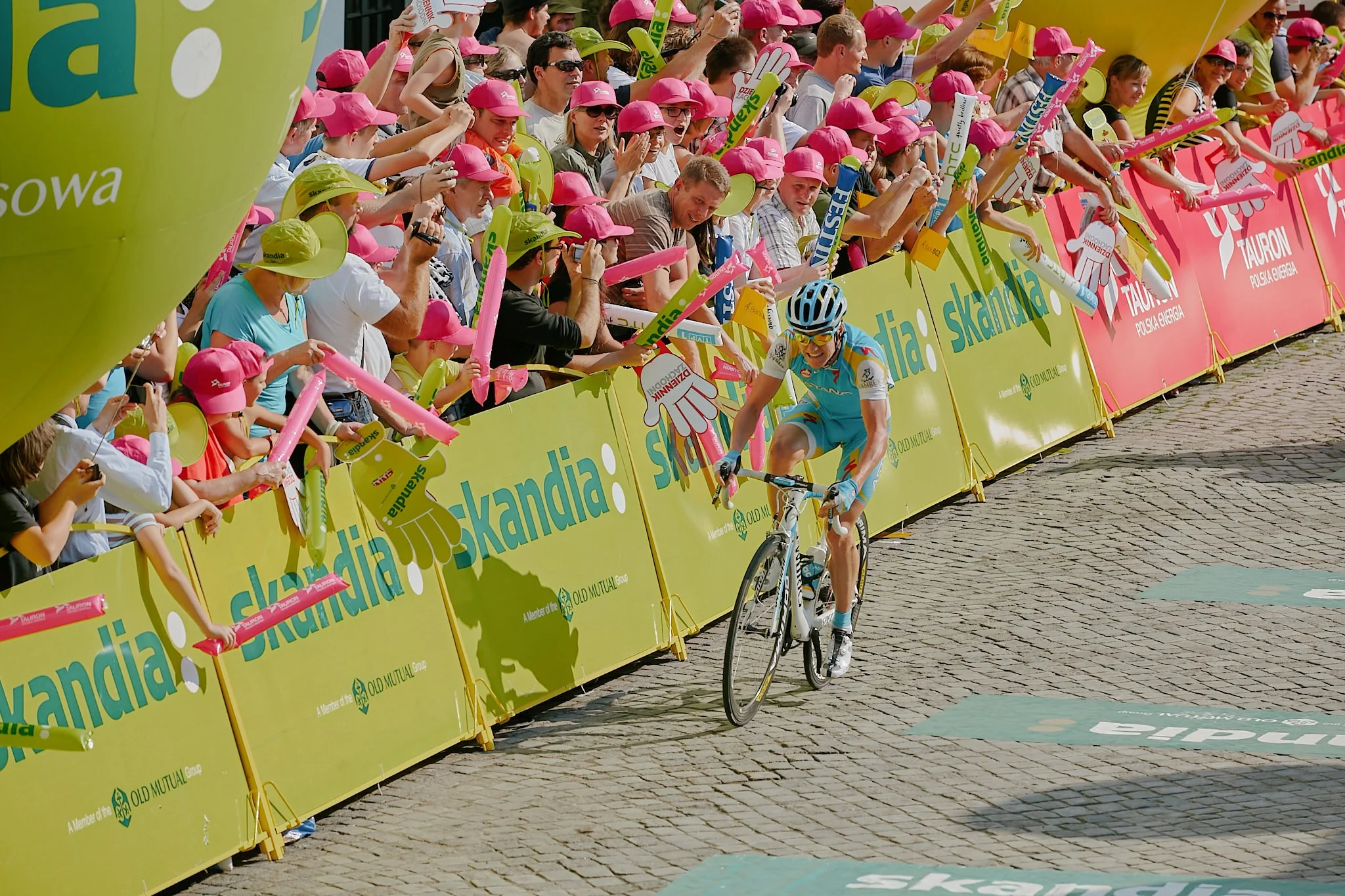 A cyclist wearing a blue and white jersey and a helmet crosses the finish line while spectators, many wearing pink and yellow hats, cheer and reach out from the side of the race track.