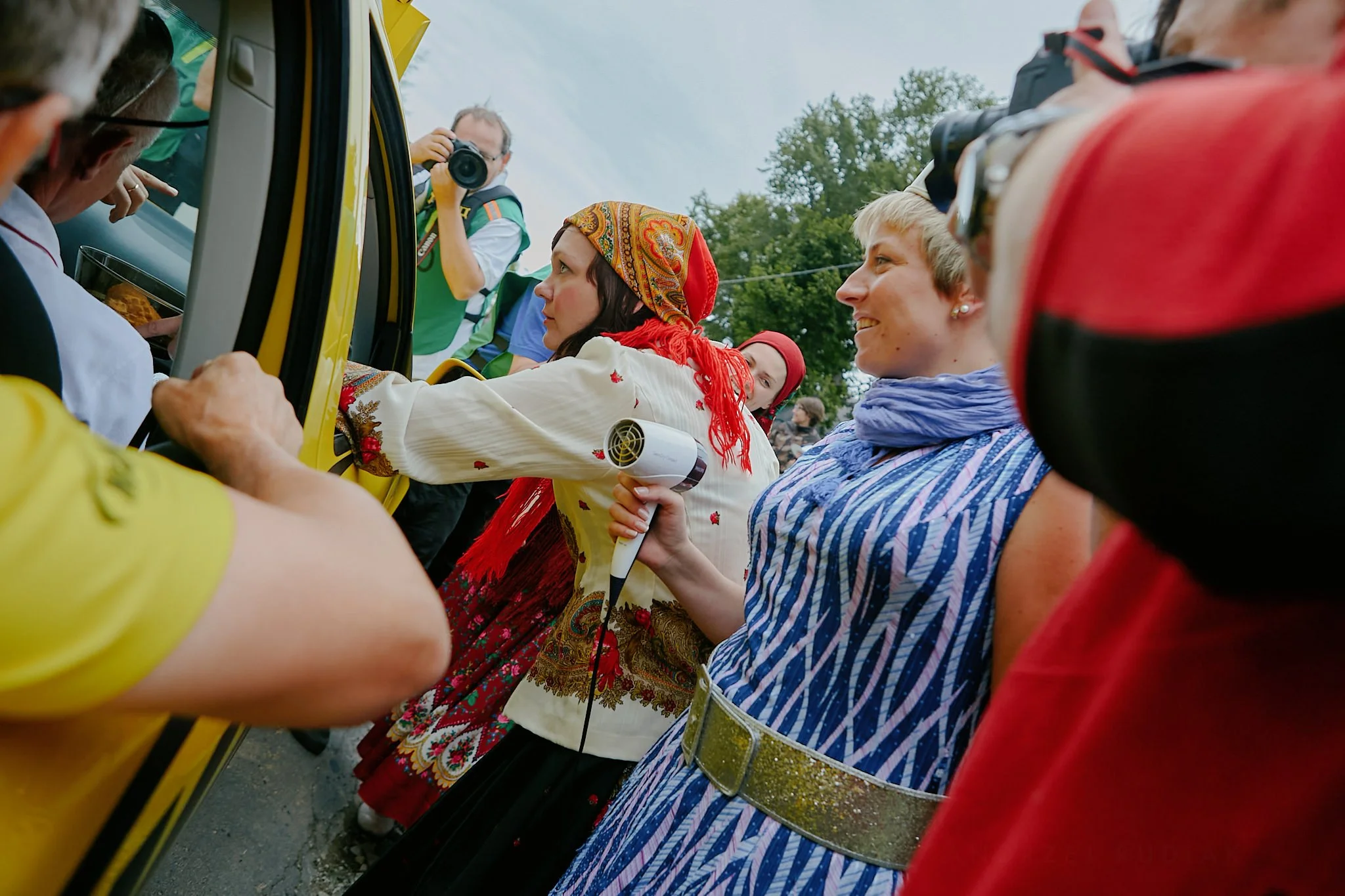 People gathered around a vehicle, with a woman dressed in traditional clothing pointing inside, and a man taking a photo. Others appear to be engaged in a cultural event outdoors.