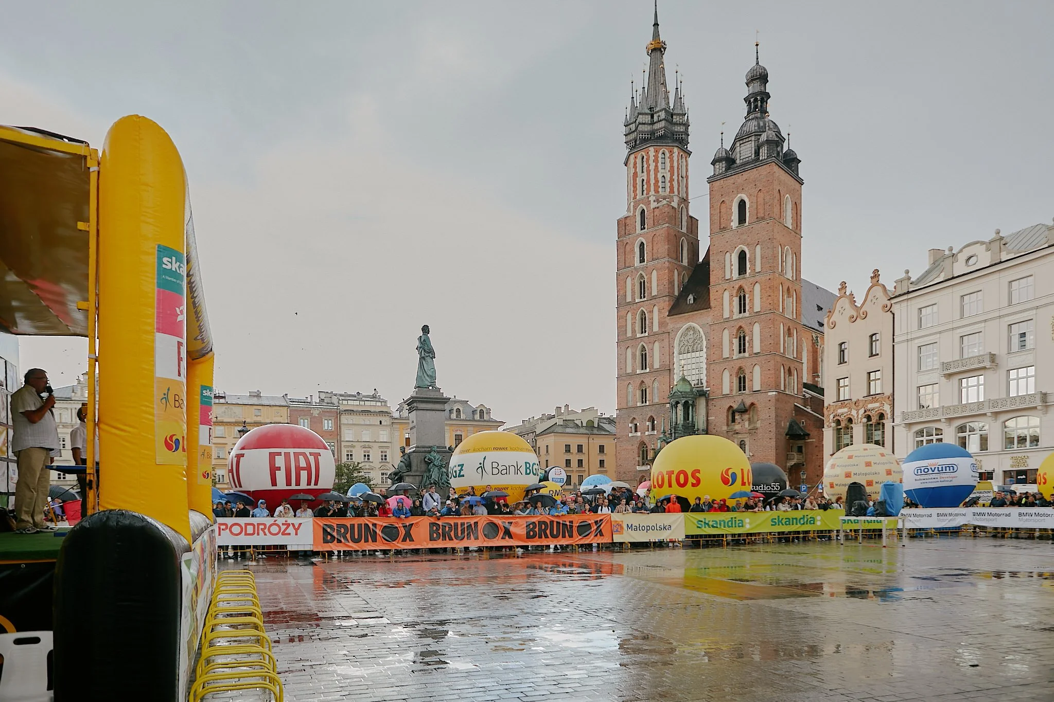 Crowd gathered in a square with colorful balloons, banners, and a large yellow inflatable structure, historic buildings with twin towers in the background, and rainy weather.
