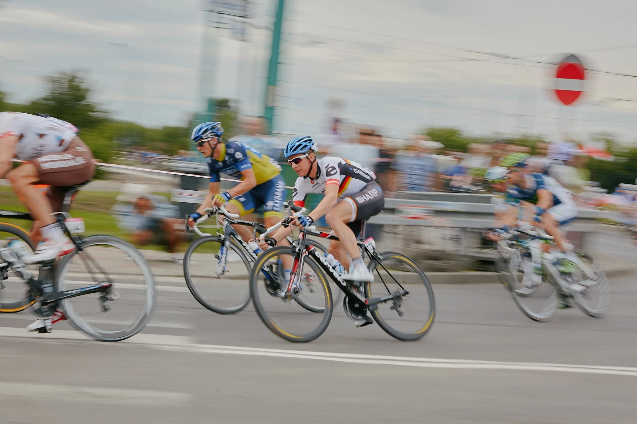 Cyclists riding bikes on a road during a race, with a red and white traffic sign and trees in the background, motion blur indicating fast movement.