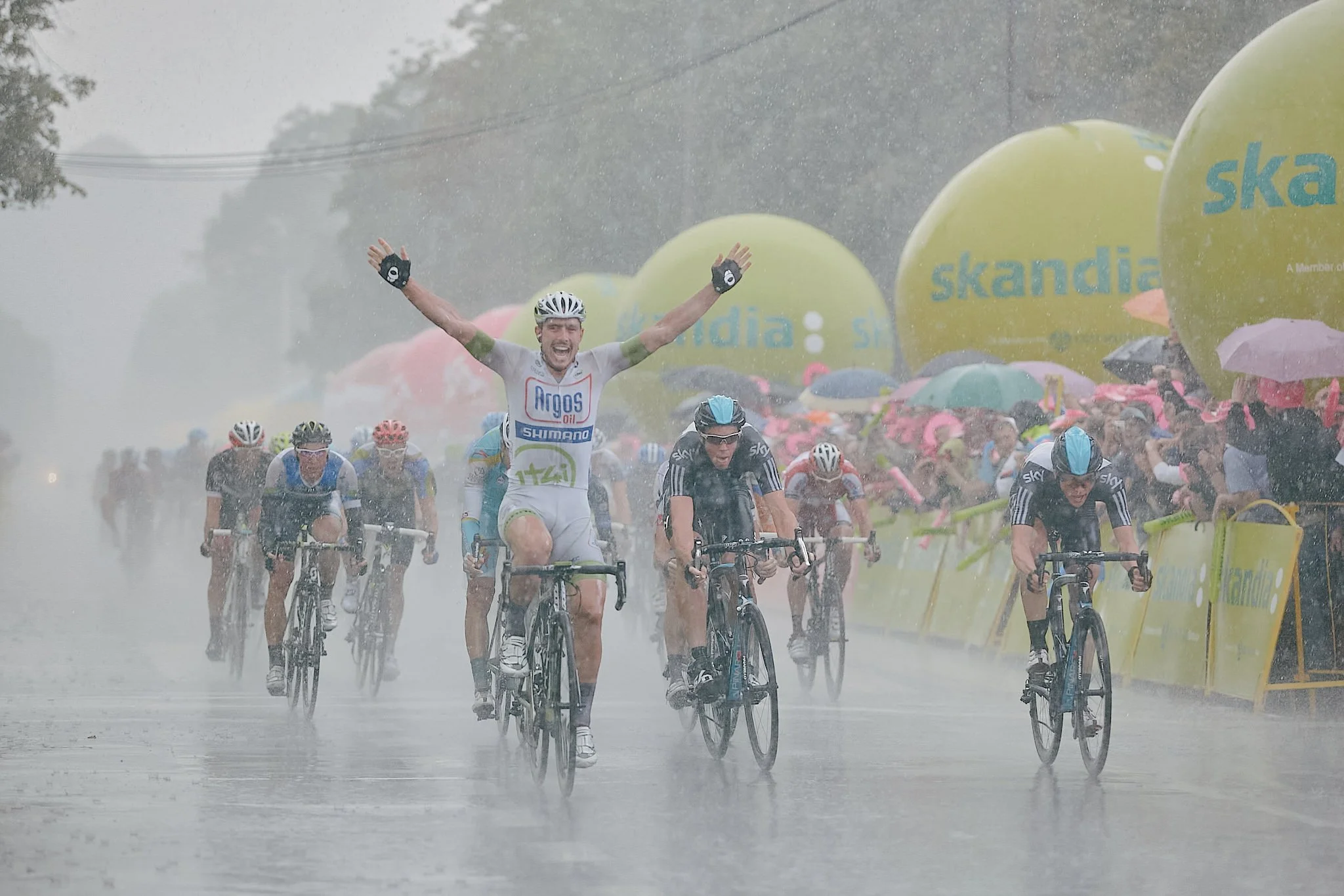 Cyclists riding their bikes in heavy rain during a race with a large crowd of spectators holding umbrellas, with yellow balloons in the background.