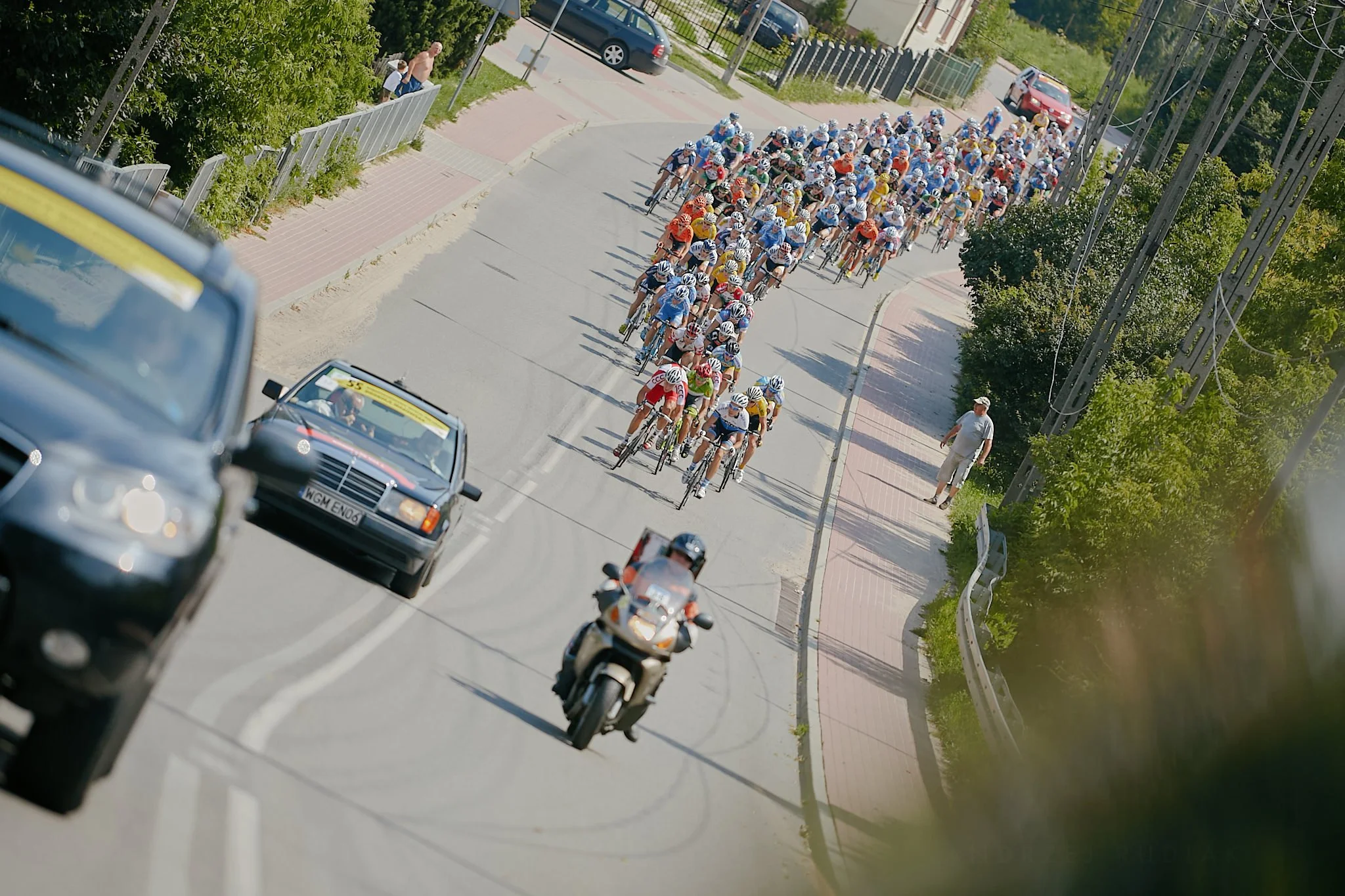 A large group of cyclists participating in a race, riding on a road lined with greenery and power lines, with a few cars and a police motorcycle leading the pack.