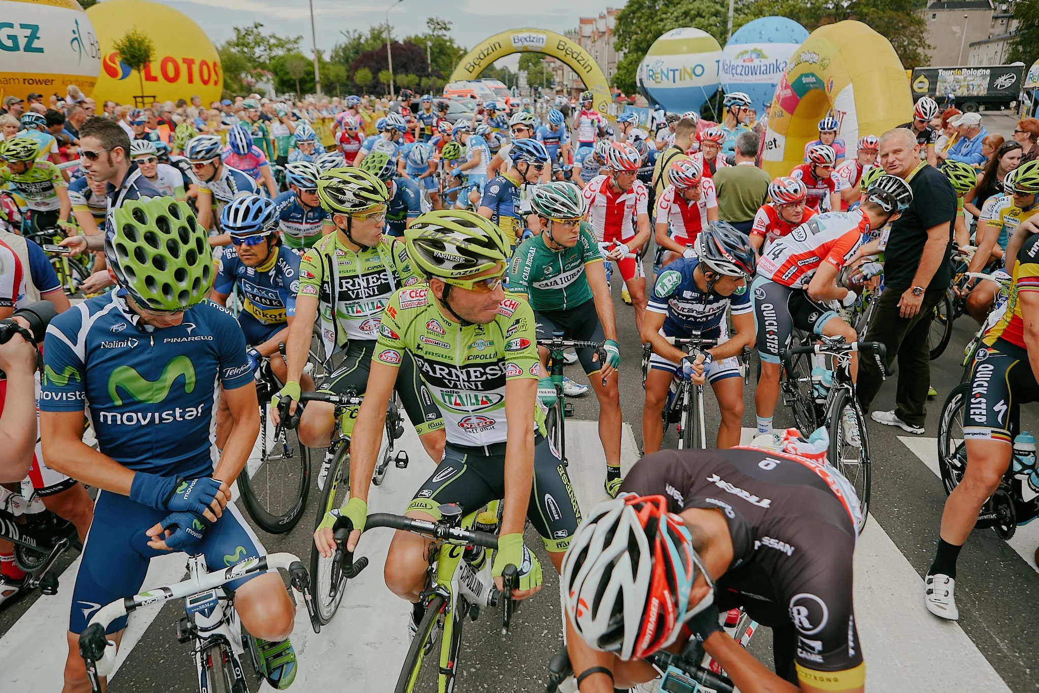 Cyclists gathered at the starting line for a race, wearing helmets and colorful racing uniforms, with spectators and sponsor banners in the background.