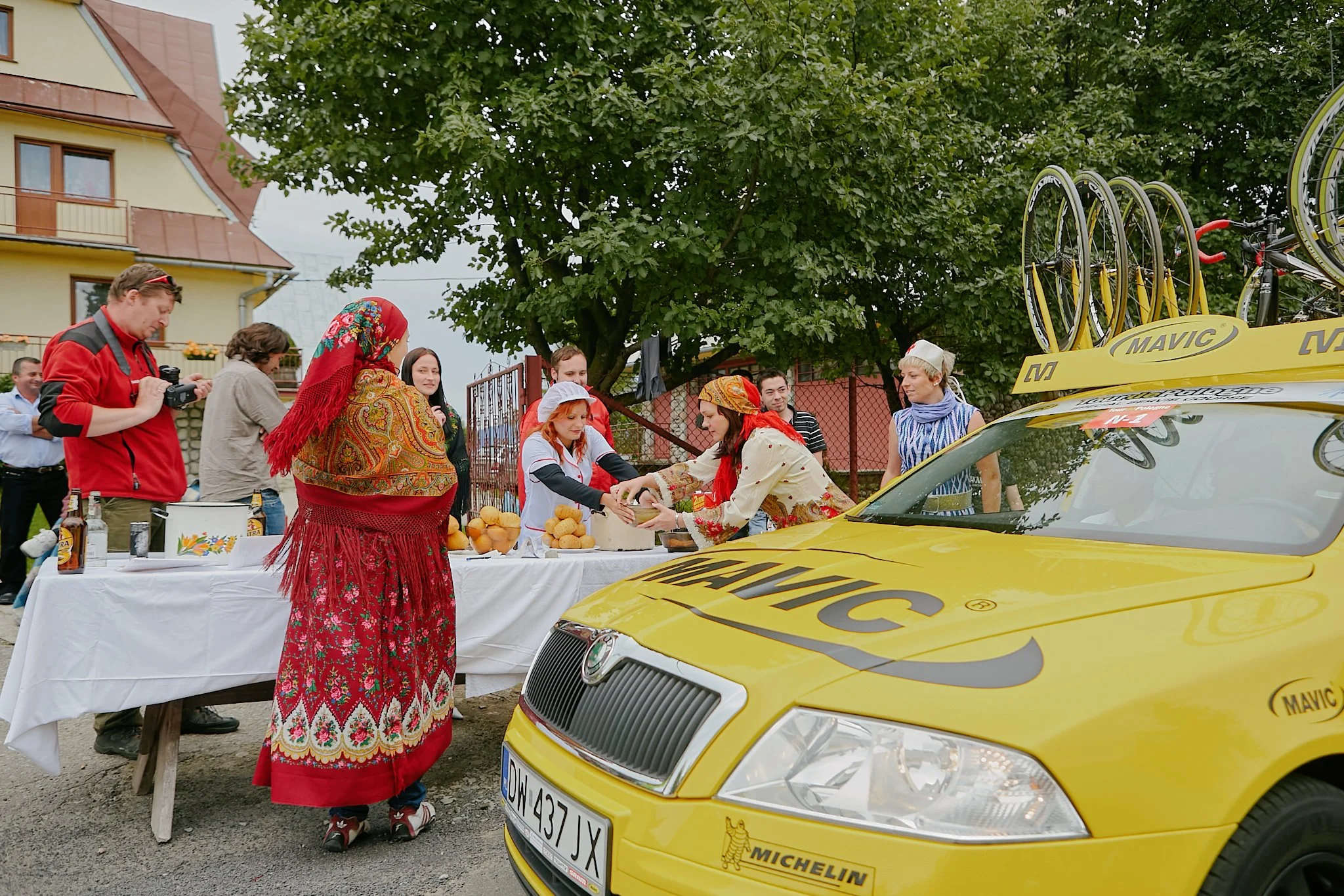 A group of people dressed in traditional Eastern European clothing gathered around a table with bread, oranges, and drinks, with a yellow race car marked with Michelin branding and spare bicycles on the roof, outdoors near a building and a large tree