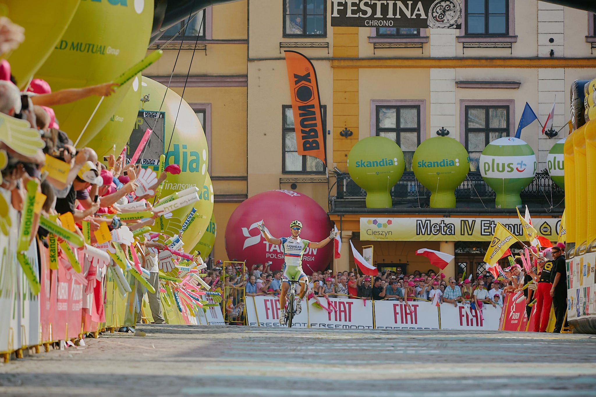 A cyclist crossing the finish line at a race event, surrounded by cheering spectators, colorful balloons, and banners in an urban setting.