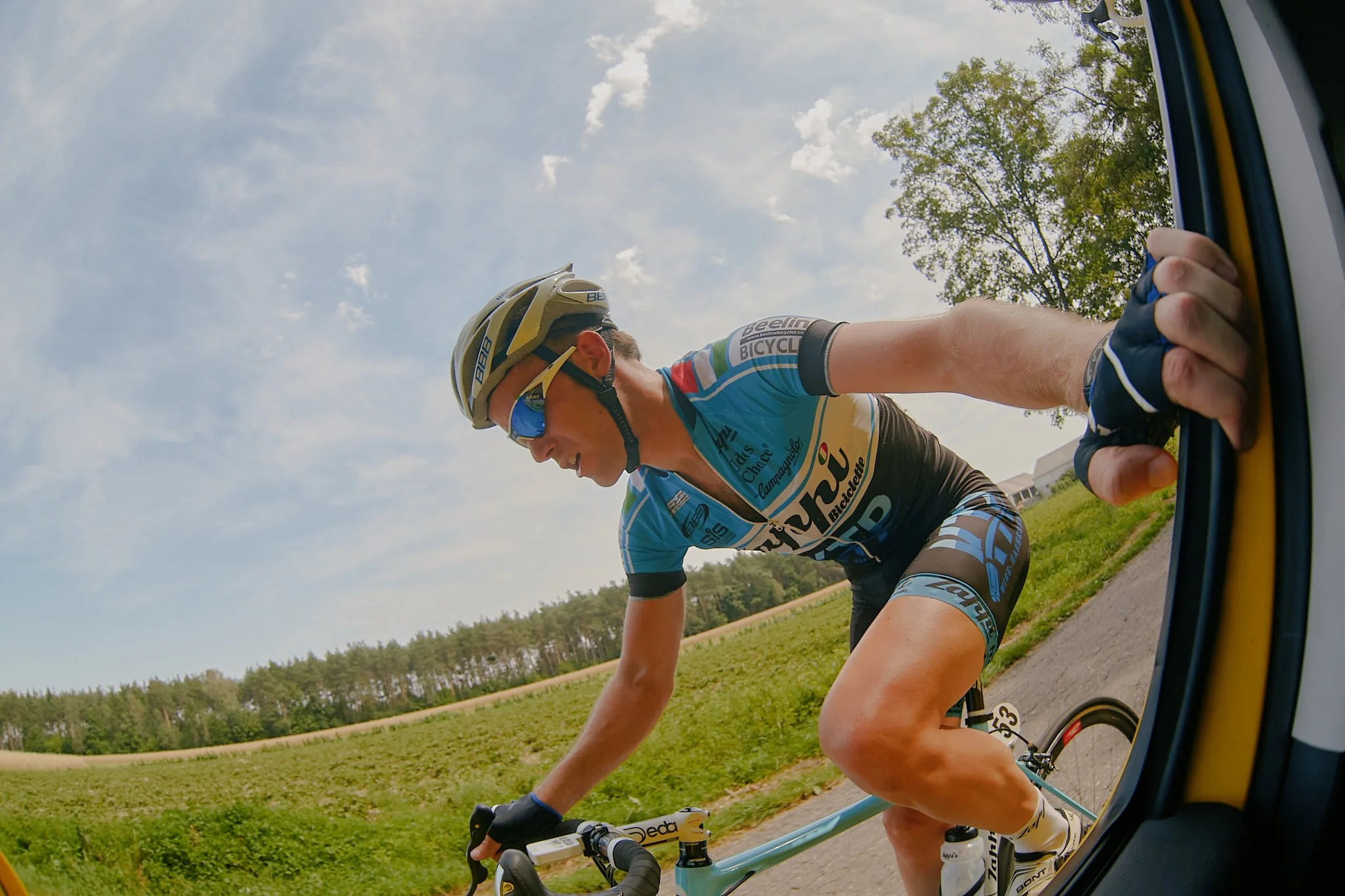 Cyclist wearing a helmet and sunglasses riding a bike on a rural road with green fields and trees in the background, viewed from a vehicle window.