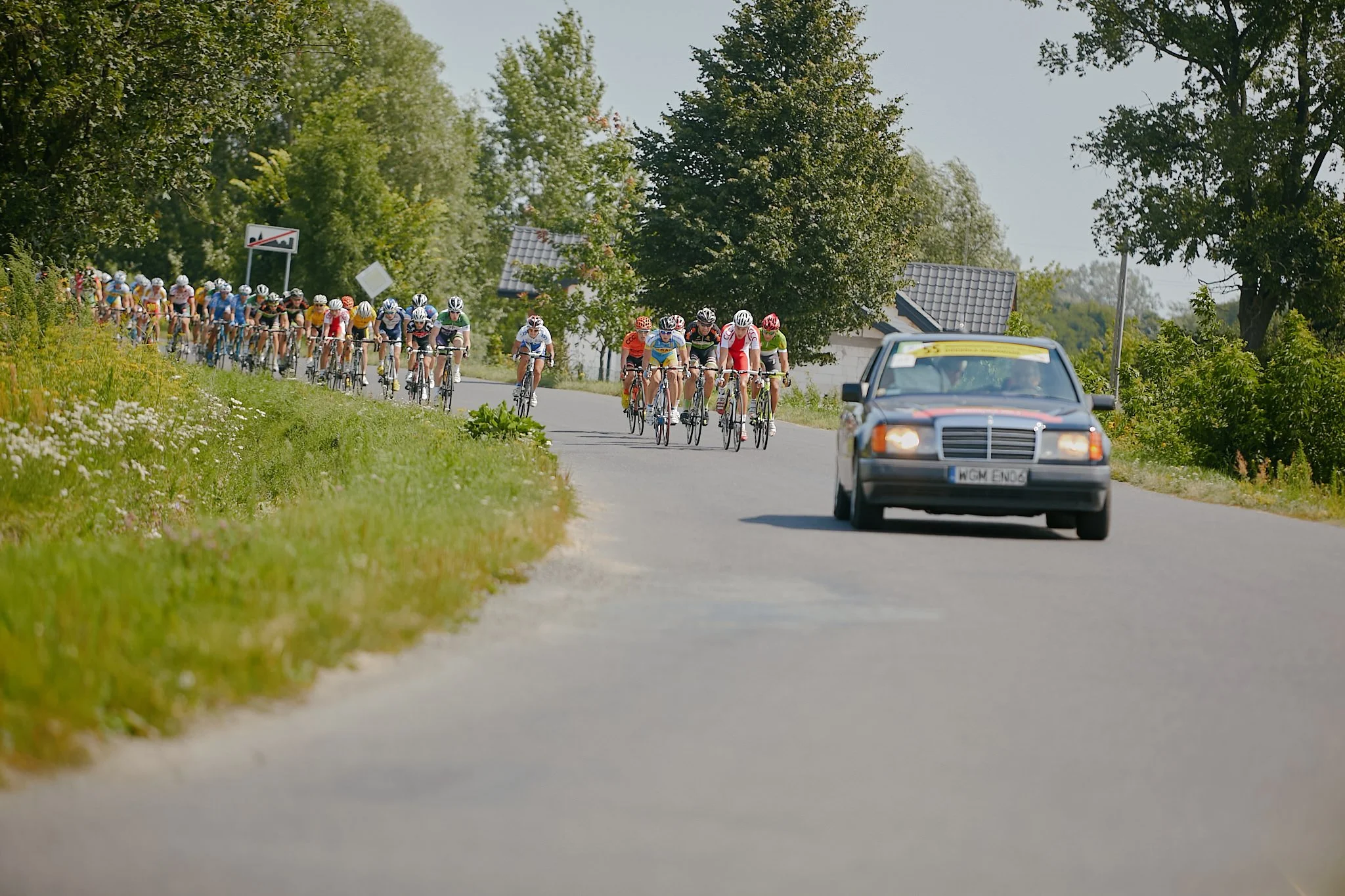 Group of cyclists participating in a race on a road surrounded by trees, with a support vehicle following behind.