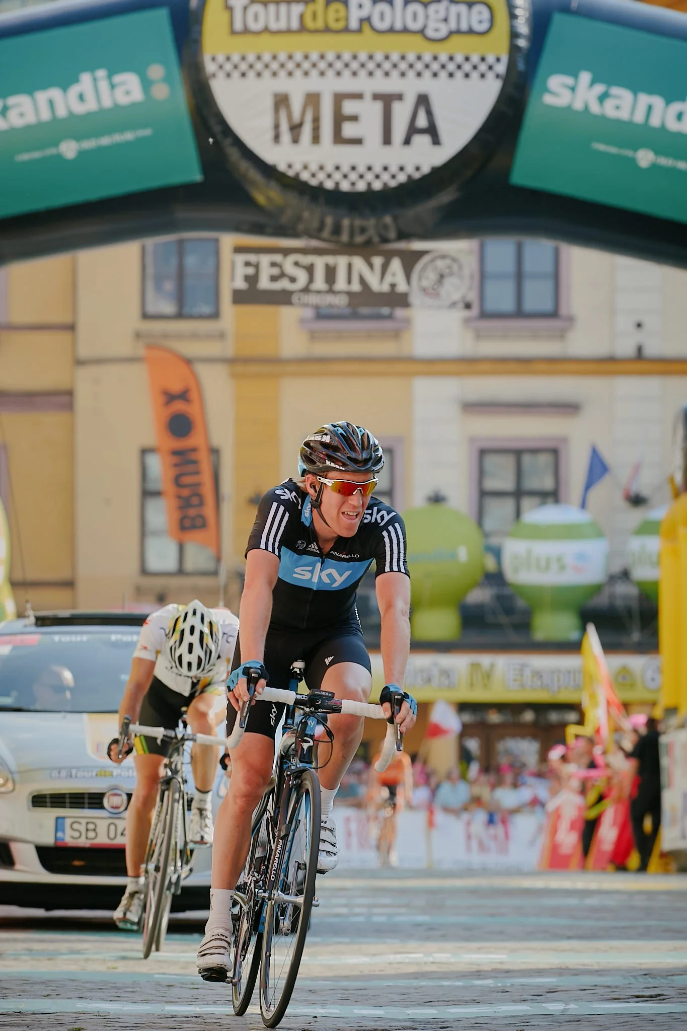Cyclists participating in a race with spectators, banners, and a town building in the background.