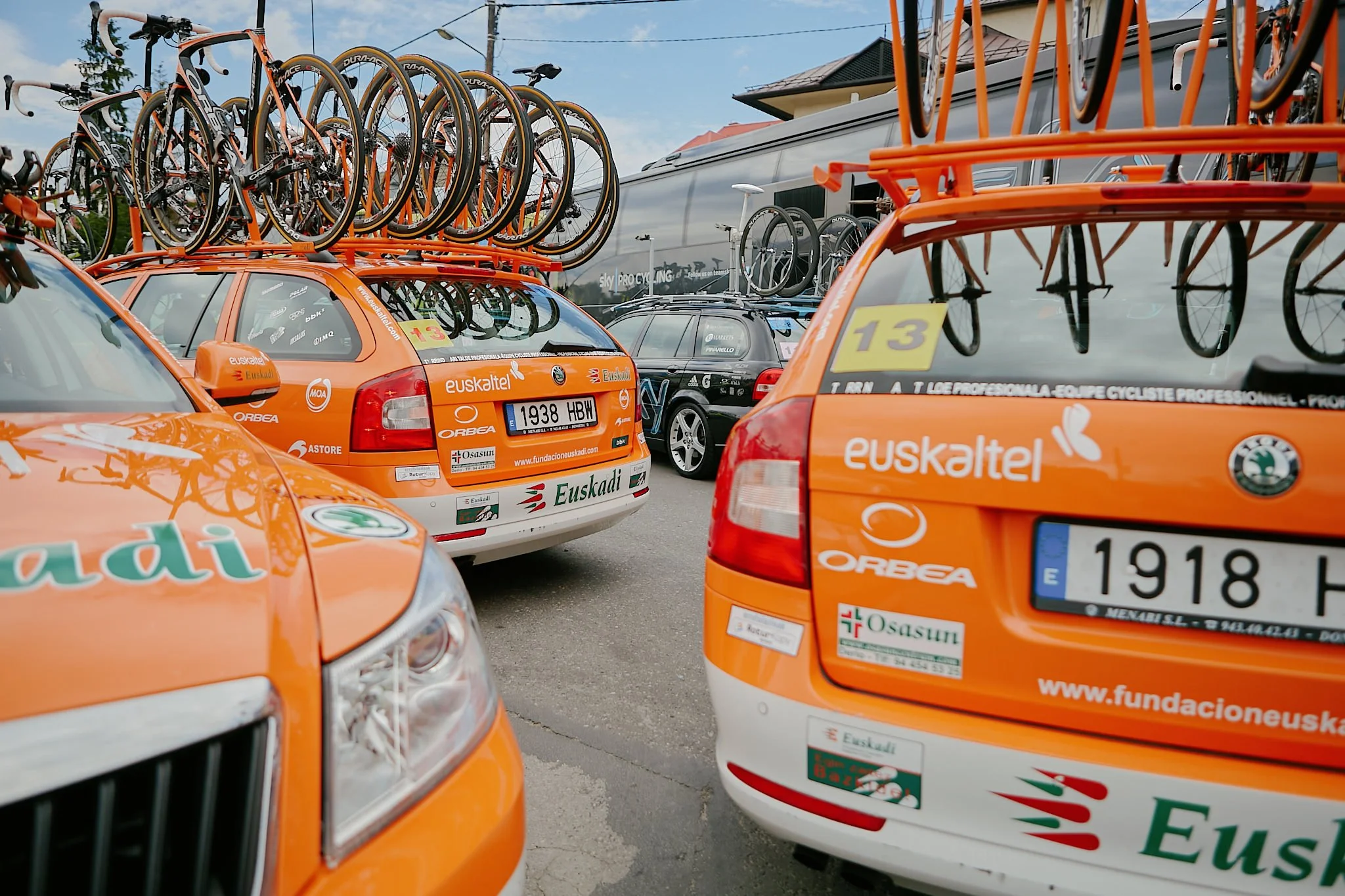 Multiple support vehicles carrying bicycles on their roofs, possibly for a cycling event or race, parked in an outdoor area with a building and cloudy sky in the background.