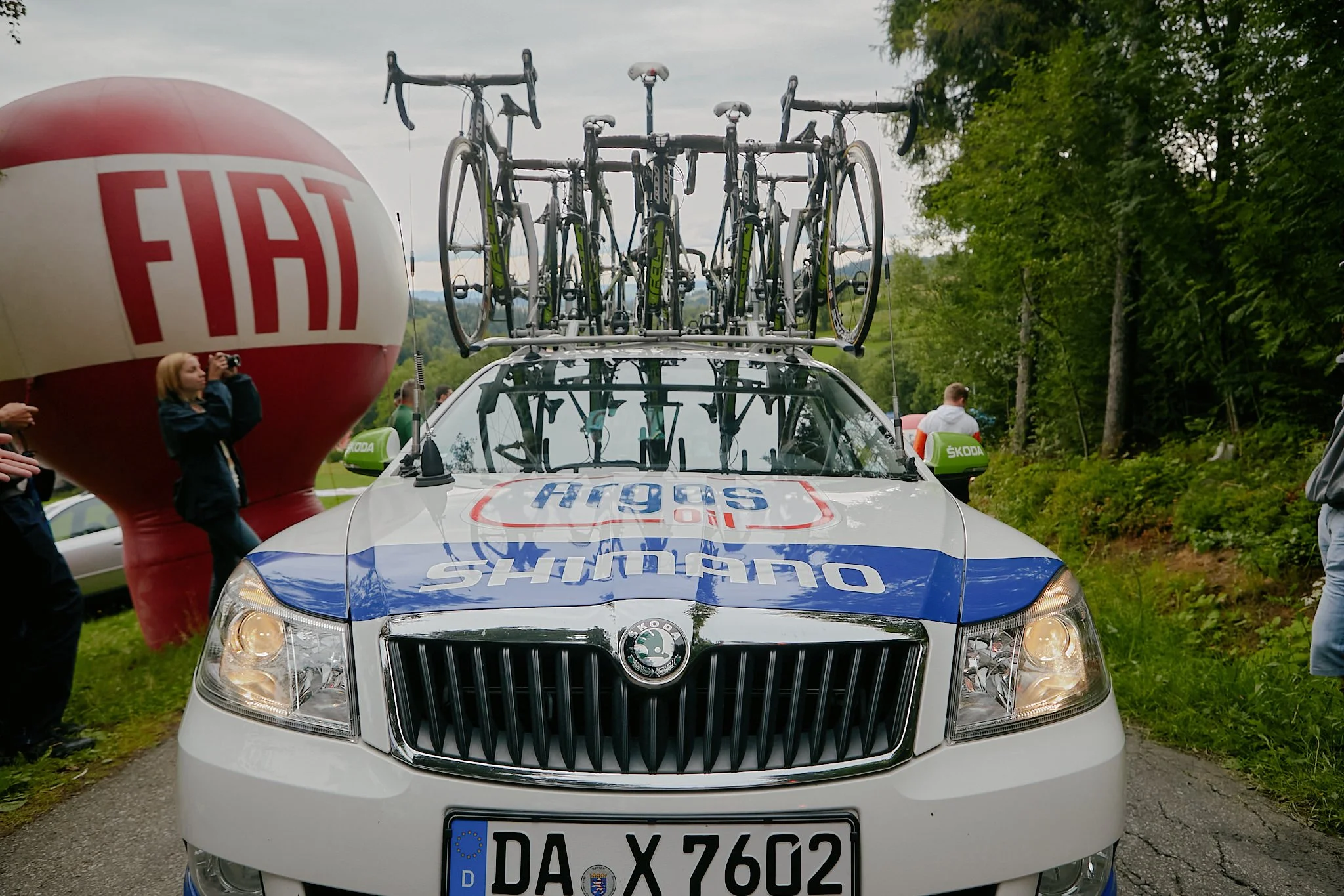 A race support vehicle with cycling race bikes mounted on top is parked on a road, with a large red and white inflatable balloon and trees in the background.