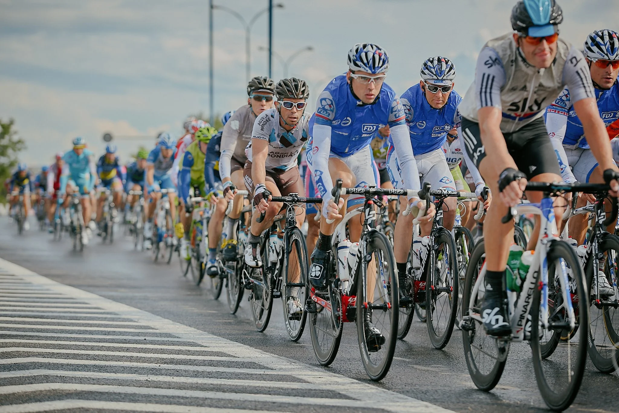 A large group of cyclists participating in a race, riding closely together on a paved road during daytime. They are wearing helmets and sports attire.