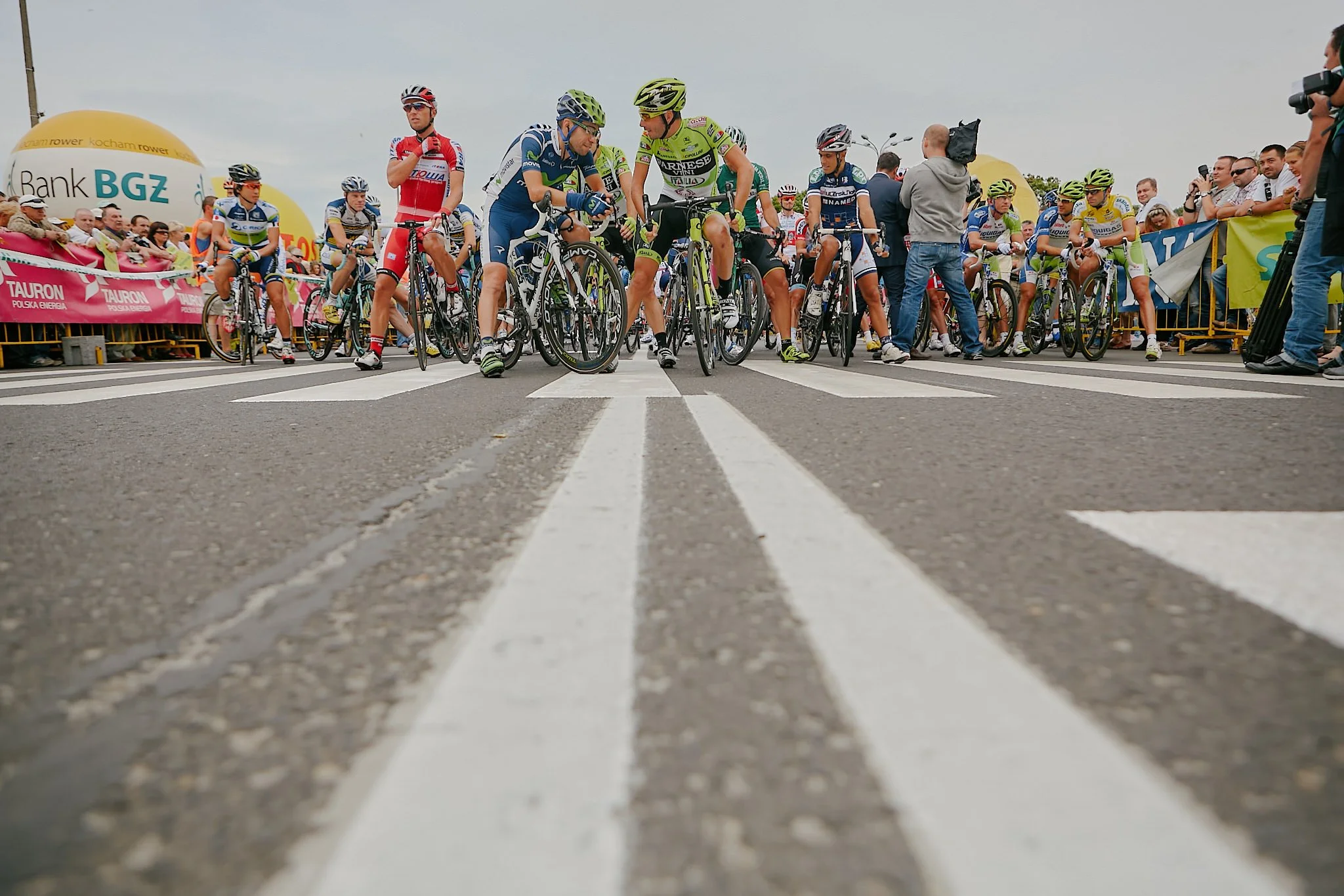 Bike race start with cyclists waiting behind a starting line, surrounded by spectators and event banners.