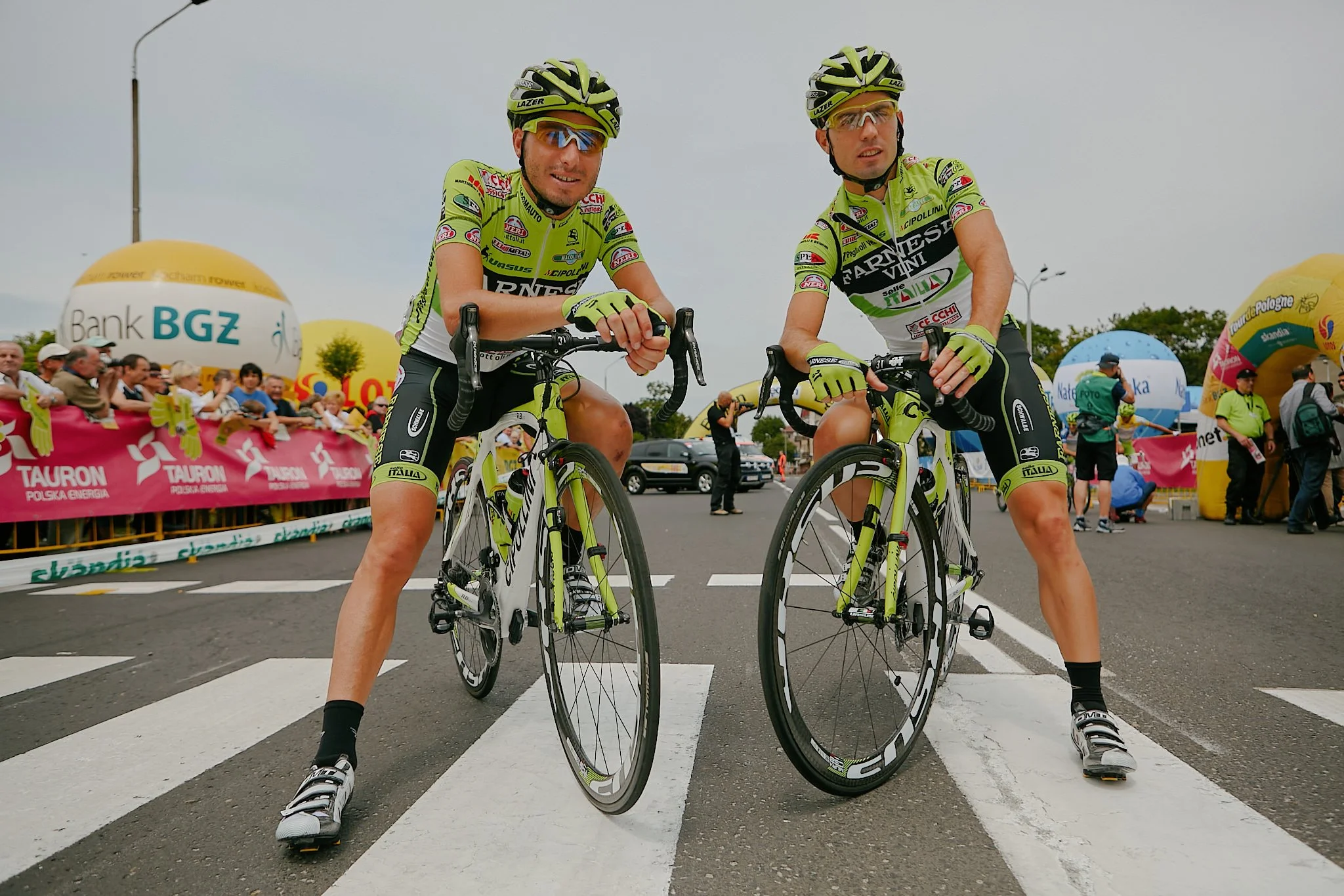 Two male cyclists in lime green jerseys and helmets standing with their bikes at a starting line during a race, with spectators and colorful balloons in the background.