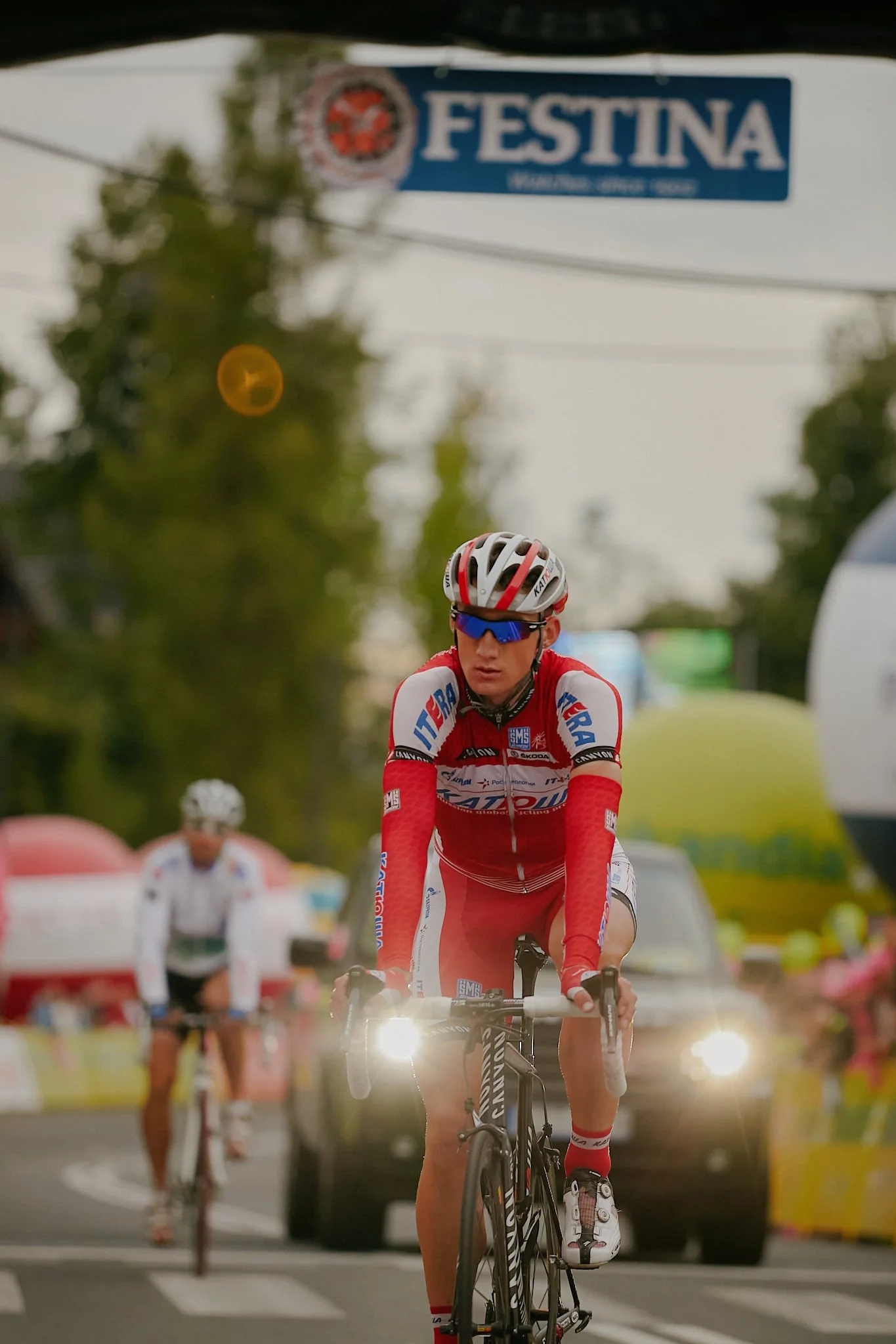 A cyclist wearing a red and white team jersey, sunglasses, and a helmet riding on a road with other cyclists and support vehicles in the background during a race.