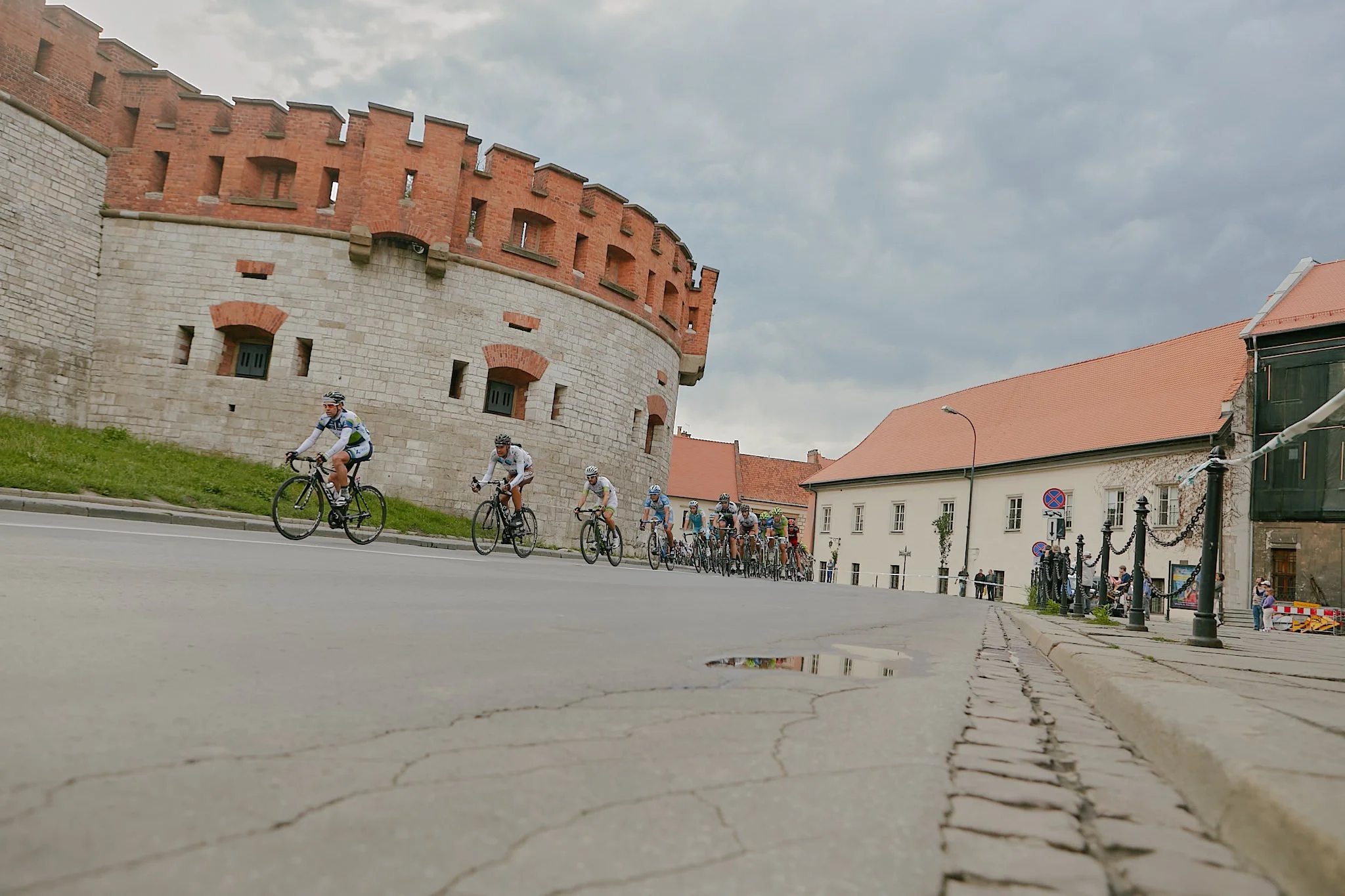 Cyclists riding in a group along a street in front of an old brick fortress and historic buildings, with cloudy sky overhead.