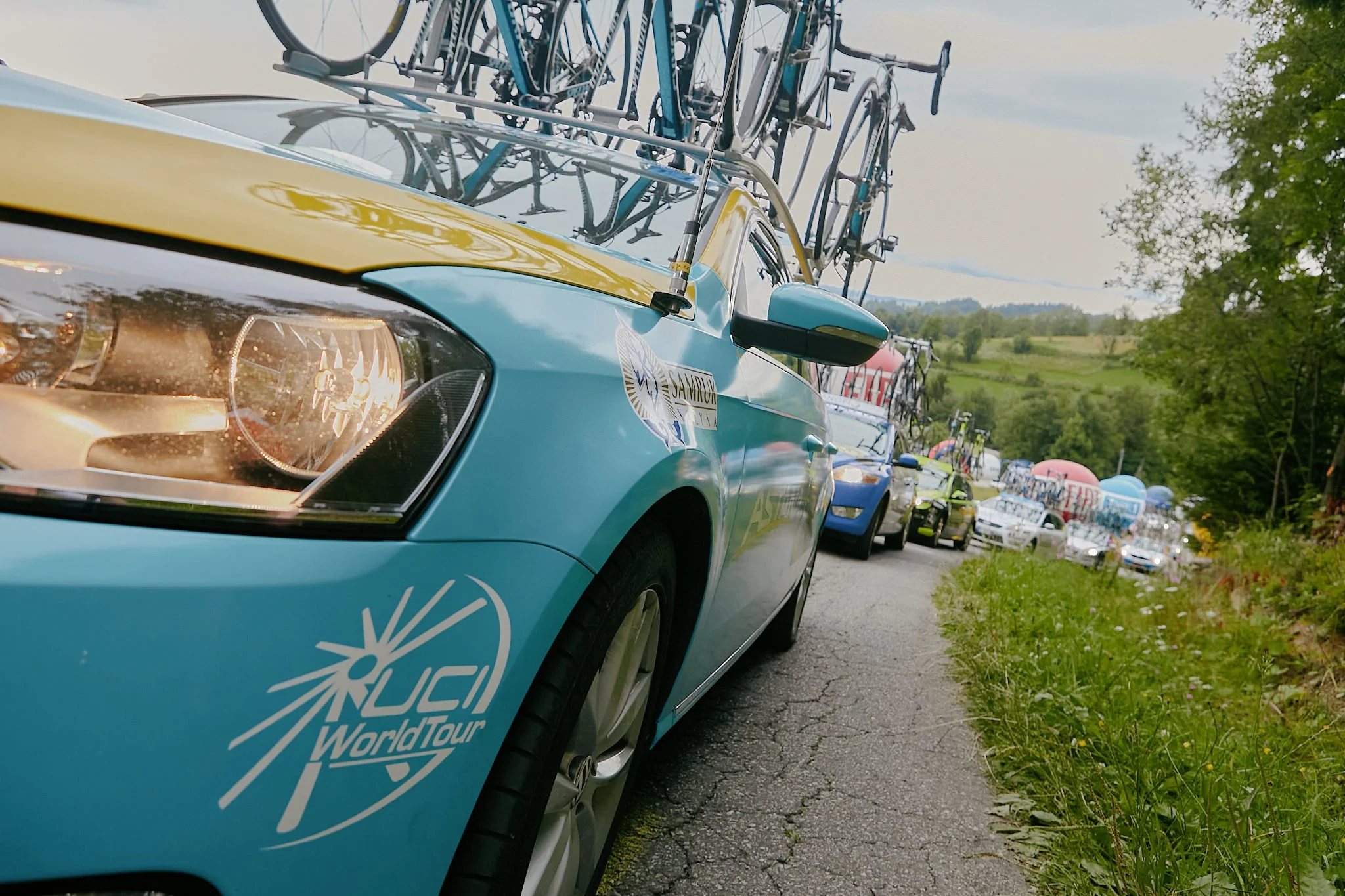 Close-up of a car with a bicycle rack mounted on the roof, carrying multiple bicycles. The car has a logo and text that reads 'WorldTour' on the side, parked on the side of a rural road with trees and a hilly landscape in the background.