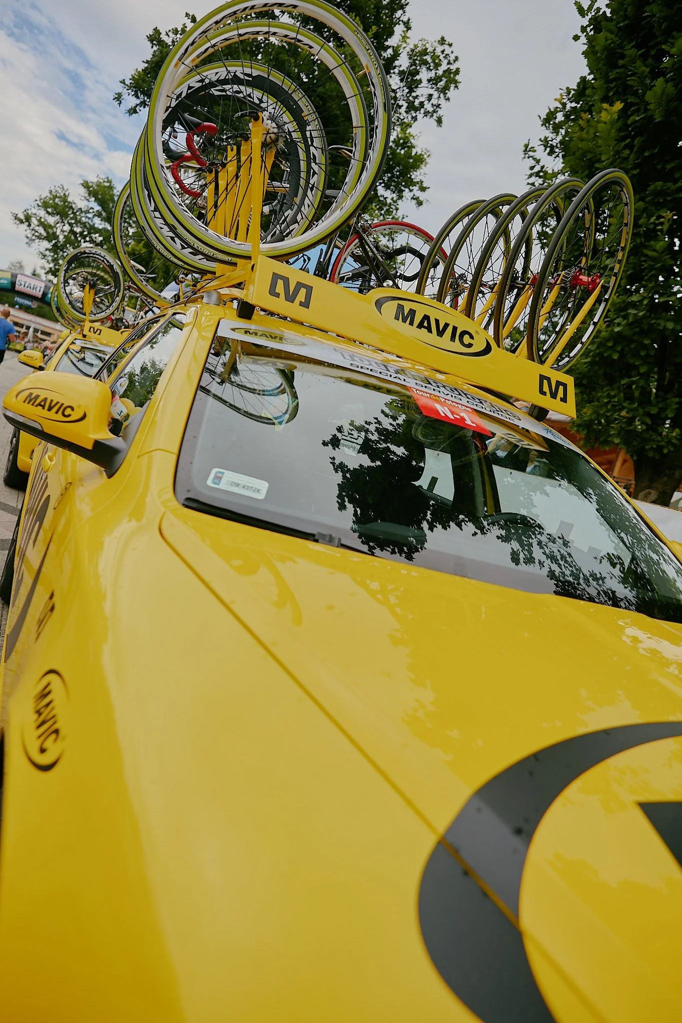 A yellow vehicle with two sets of bike racks on top, holding multiple bicycles, parked outdoors with trees and a cloudy sky in the background.