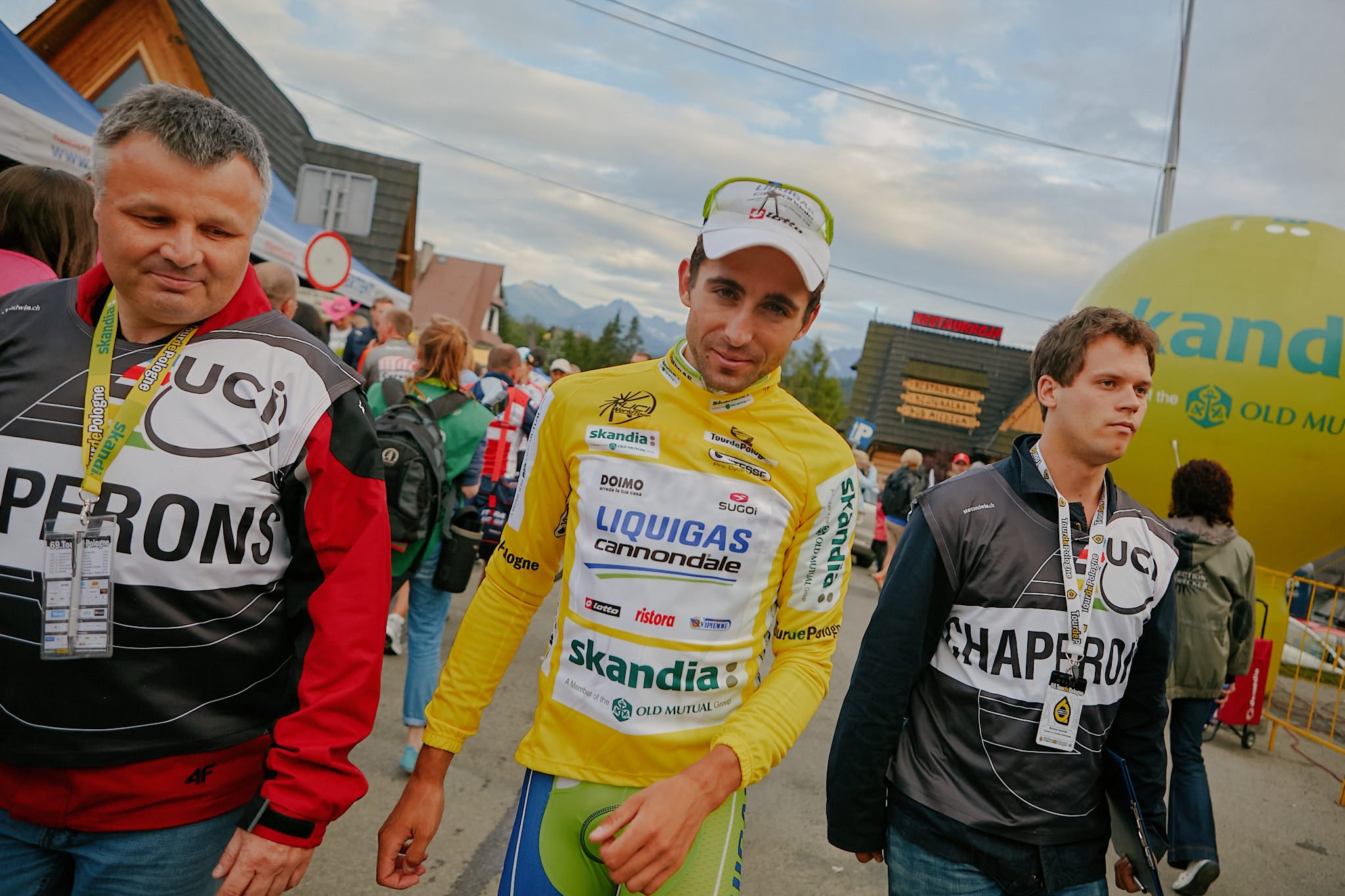 Three men are standing together outdoors, possibly at a cycling event. The man in the center is dressed in a yellow cycling jersey with various sponsor logos and is holding a pair of glasses on his head. The man on the left is wearing a black and whi