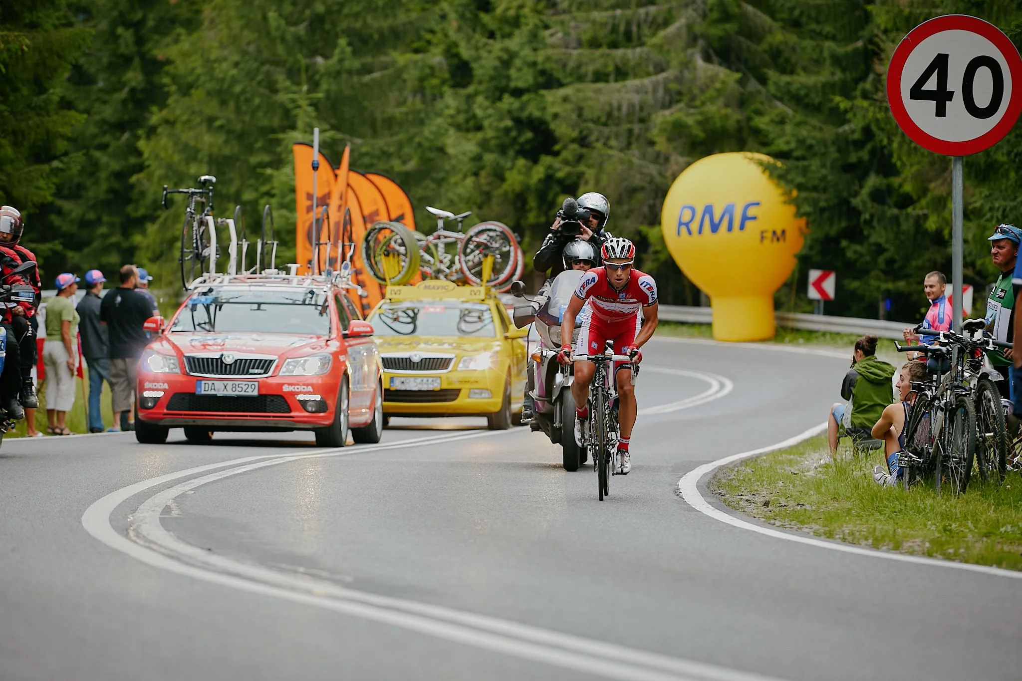 Cyclists competing in a race on a winding road, with support vehicles including a yellow car with bikes on top, a red car, and a motorcycle, surrounded by spectators and race officials.