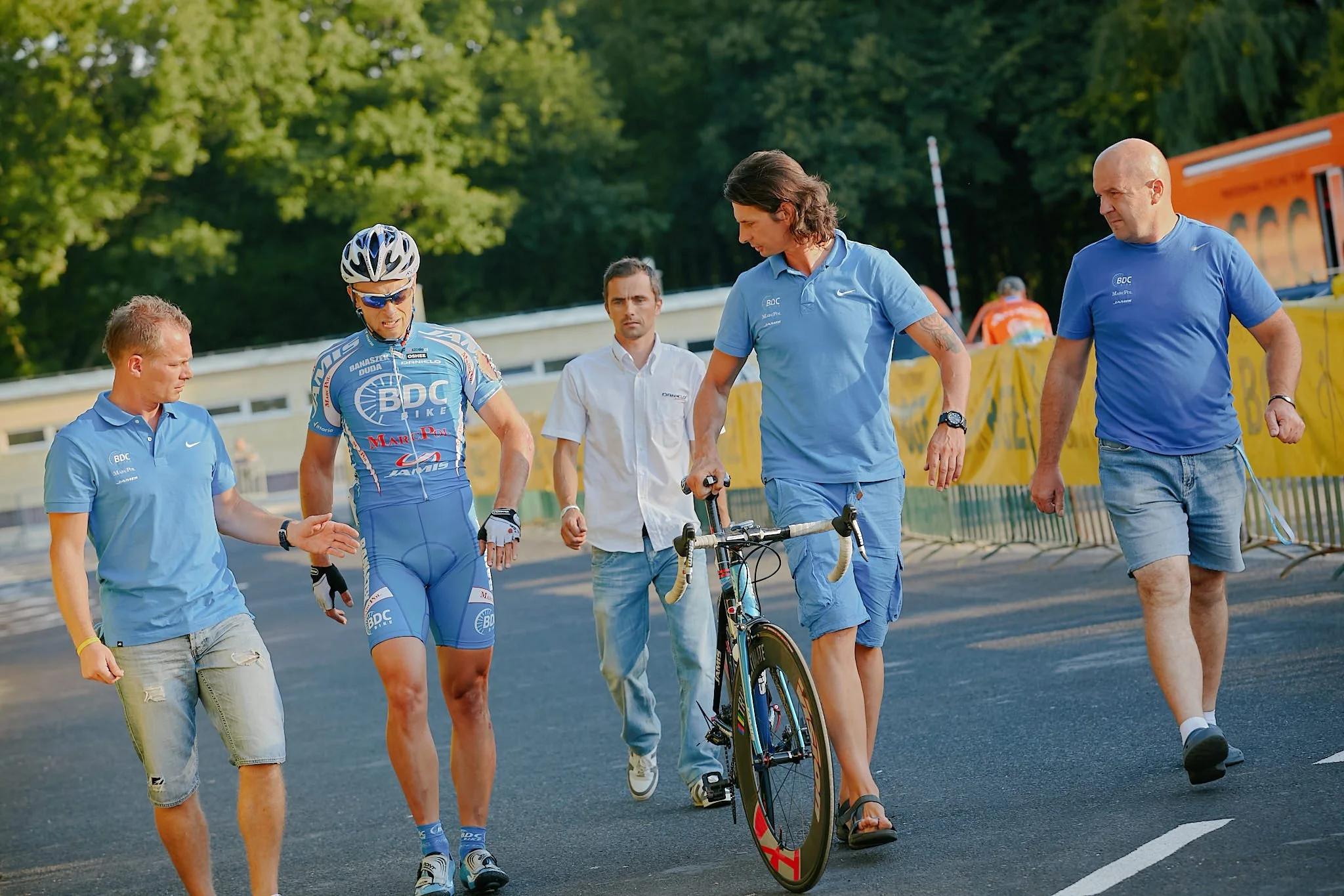 A professional cyclist in a blue jersey and helmet walking with support staff and officials, preparing to ride on a track with a teammate on a racing bicycle, surrounded by trees and event banners.