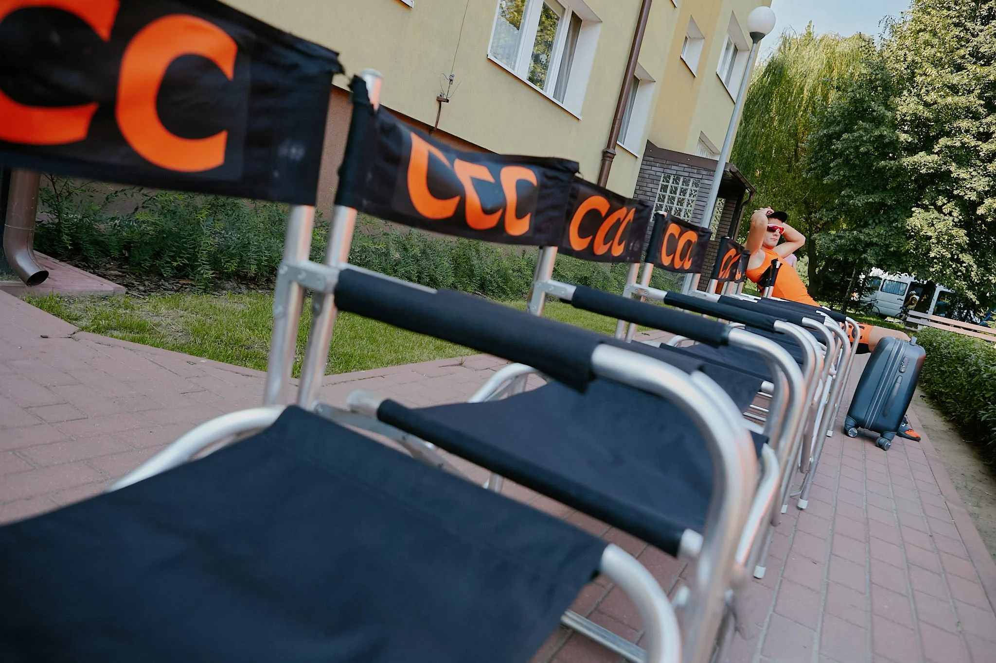 Empty lounge chairs lined up outside a building with a woman sitting with sunglasses and an orange shirt in the background, trees, and a building with windows.