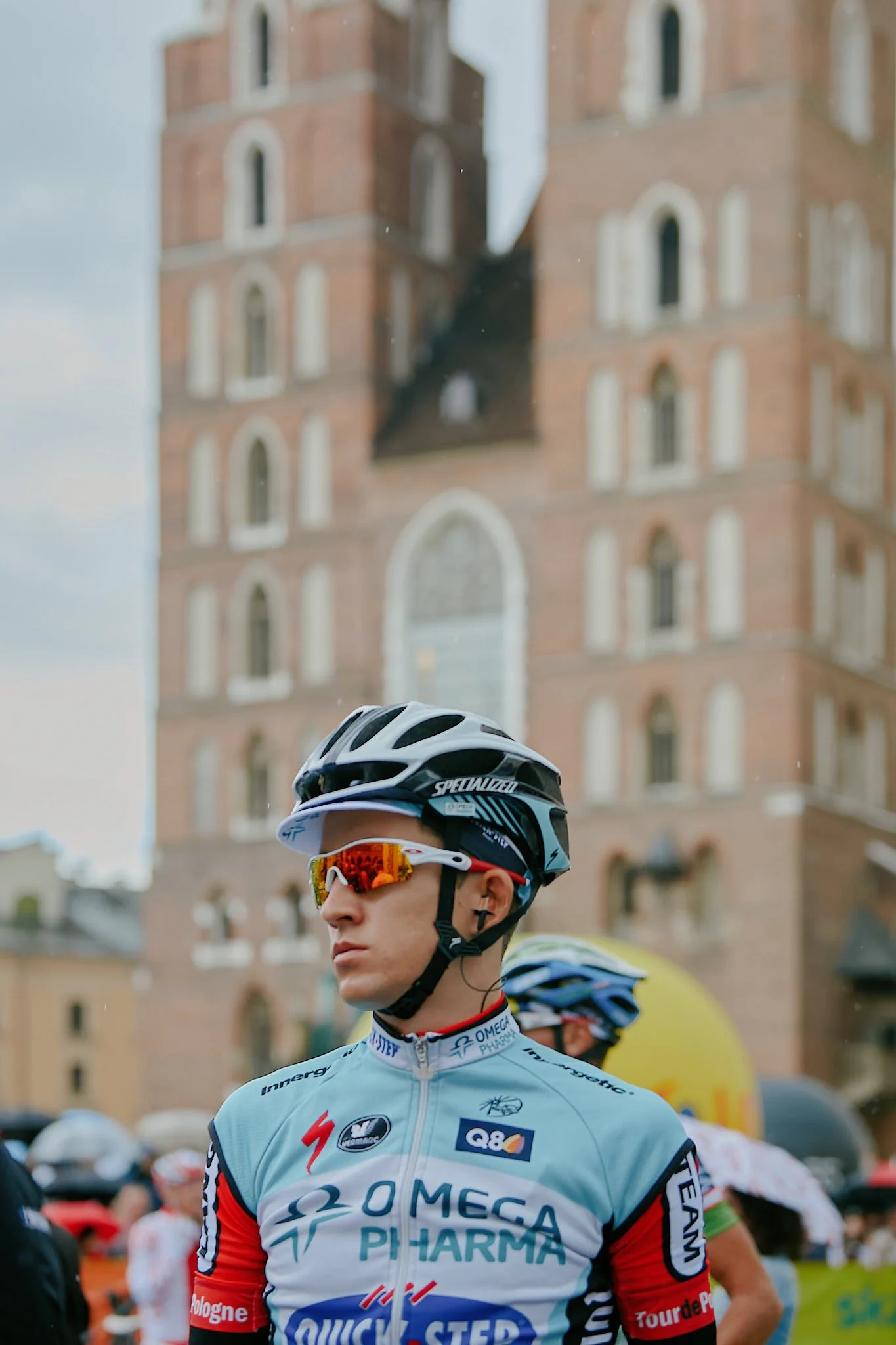 A cyclist wearing a light blue racing jersey, helmet, and sunglasses stands in front of a historic brick building with arched windows. Other cyclists and spectators are visible in the background.