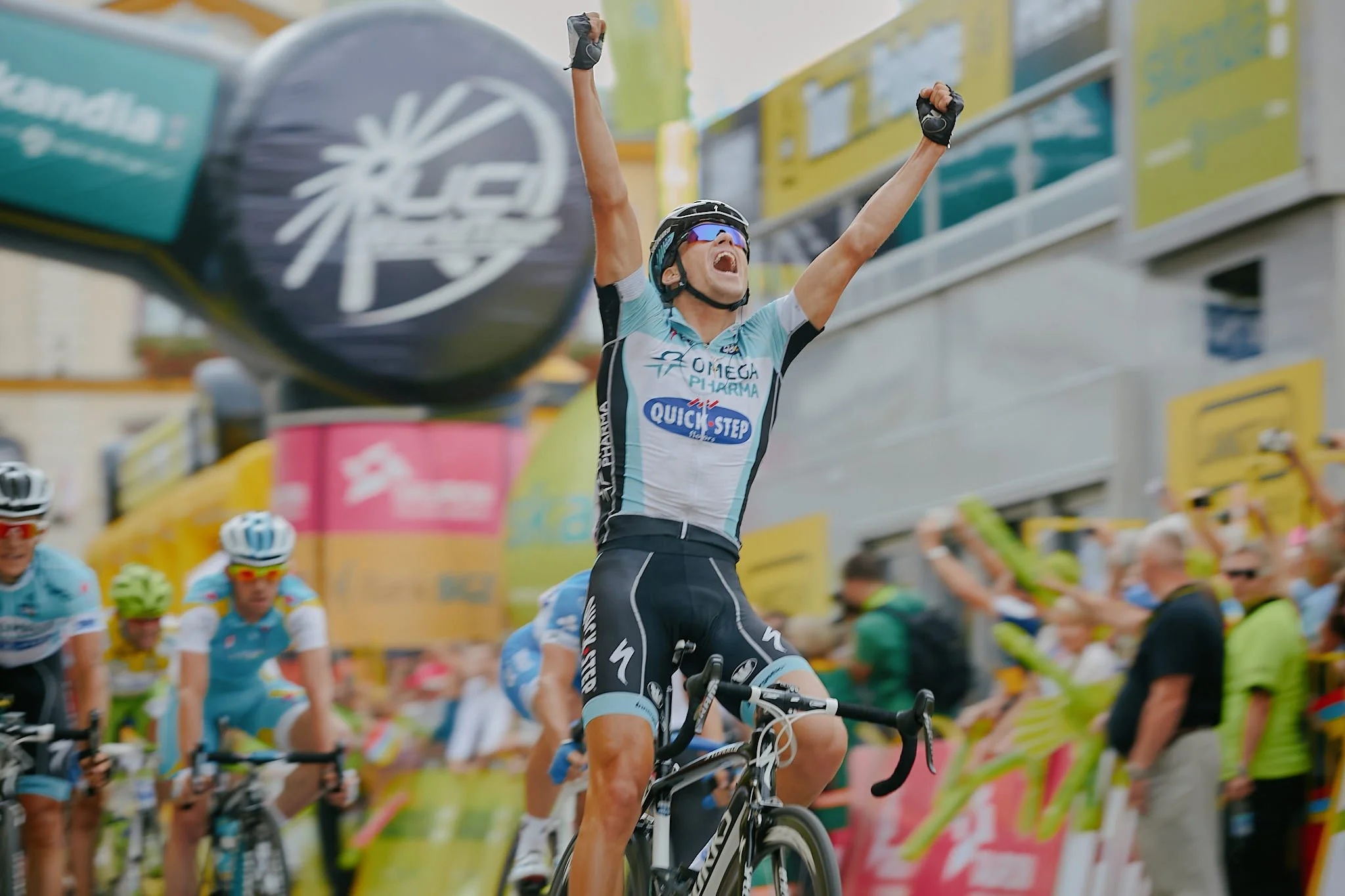 A cyclist crossing the finish line with arms raised in victory at a race, surrounded by other cyclists and spectators.
