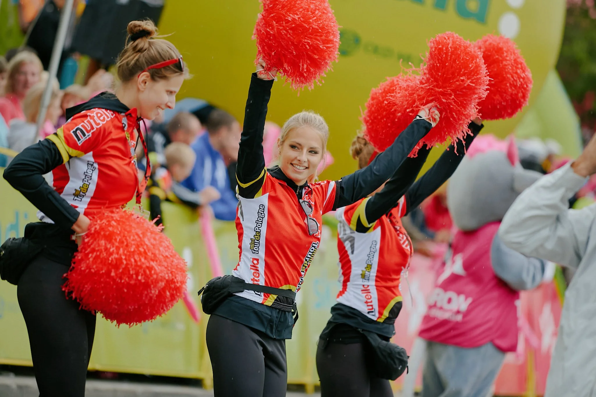 Two women in red and black cycling jerseys, smiling and holding red pom-poms during a cycling event with spectators in the background.