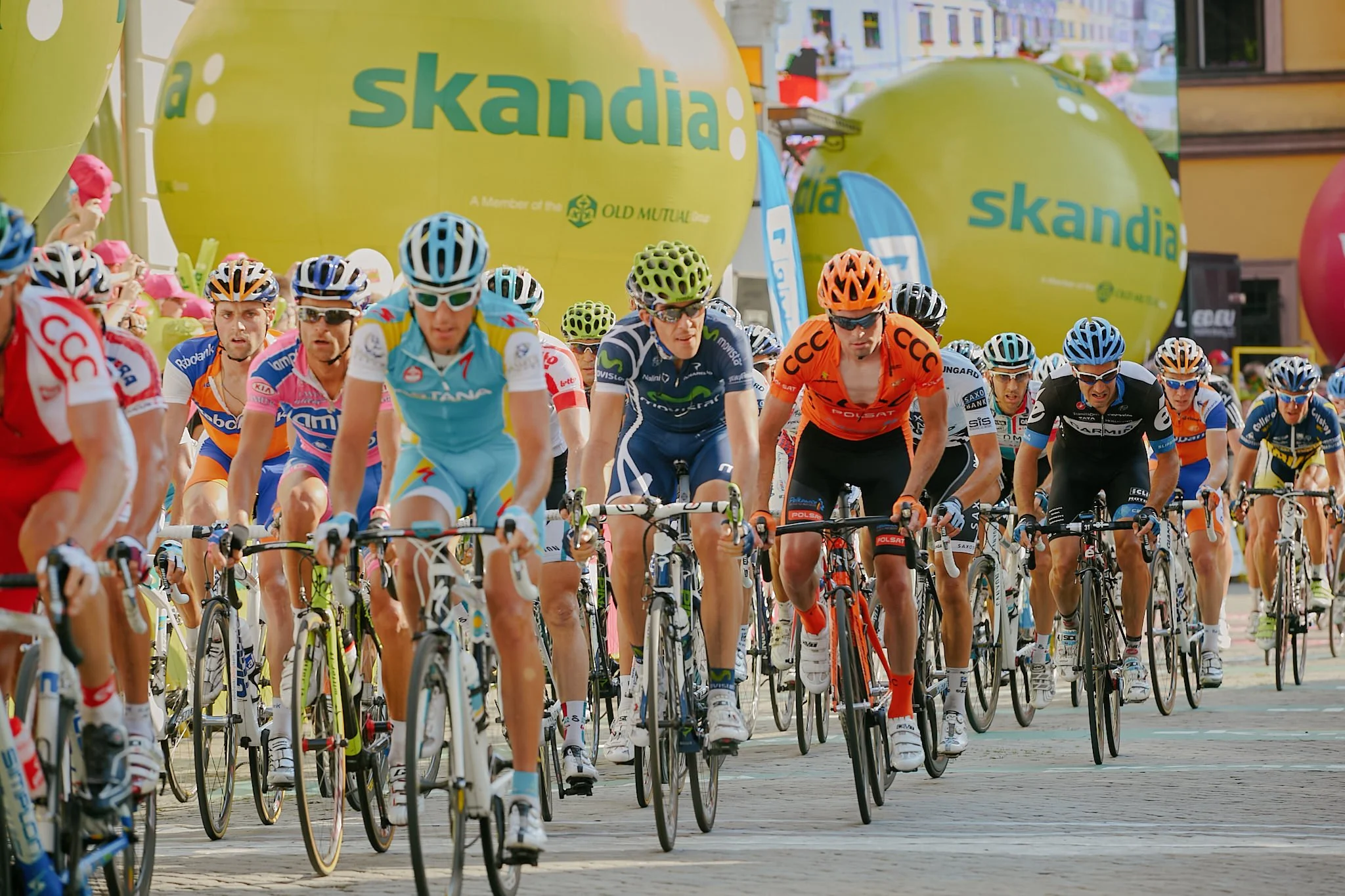 Cyclists participating in a race, wearing helmets and colorful jerseys, riding on a city street lined with large yellow balloons and banners.