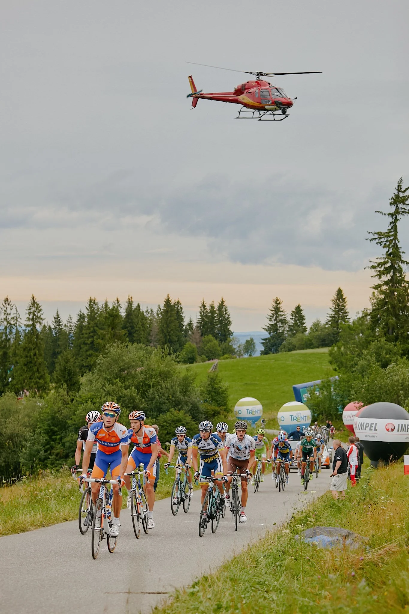 Cyclists participating in a race on a rural road, with spectators and event banners along the sides, and a helicopter flying overhead.