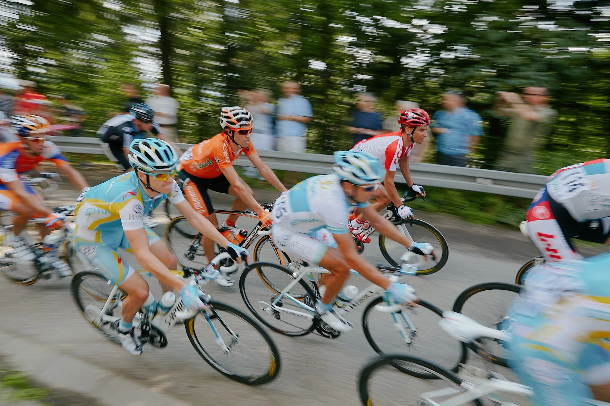 Group of cyclists racing on a paved road, wearing helmets and colorful sports attire, with motion blur indicating their fast movement, in a lush green outdoor setting.