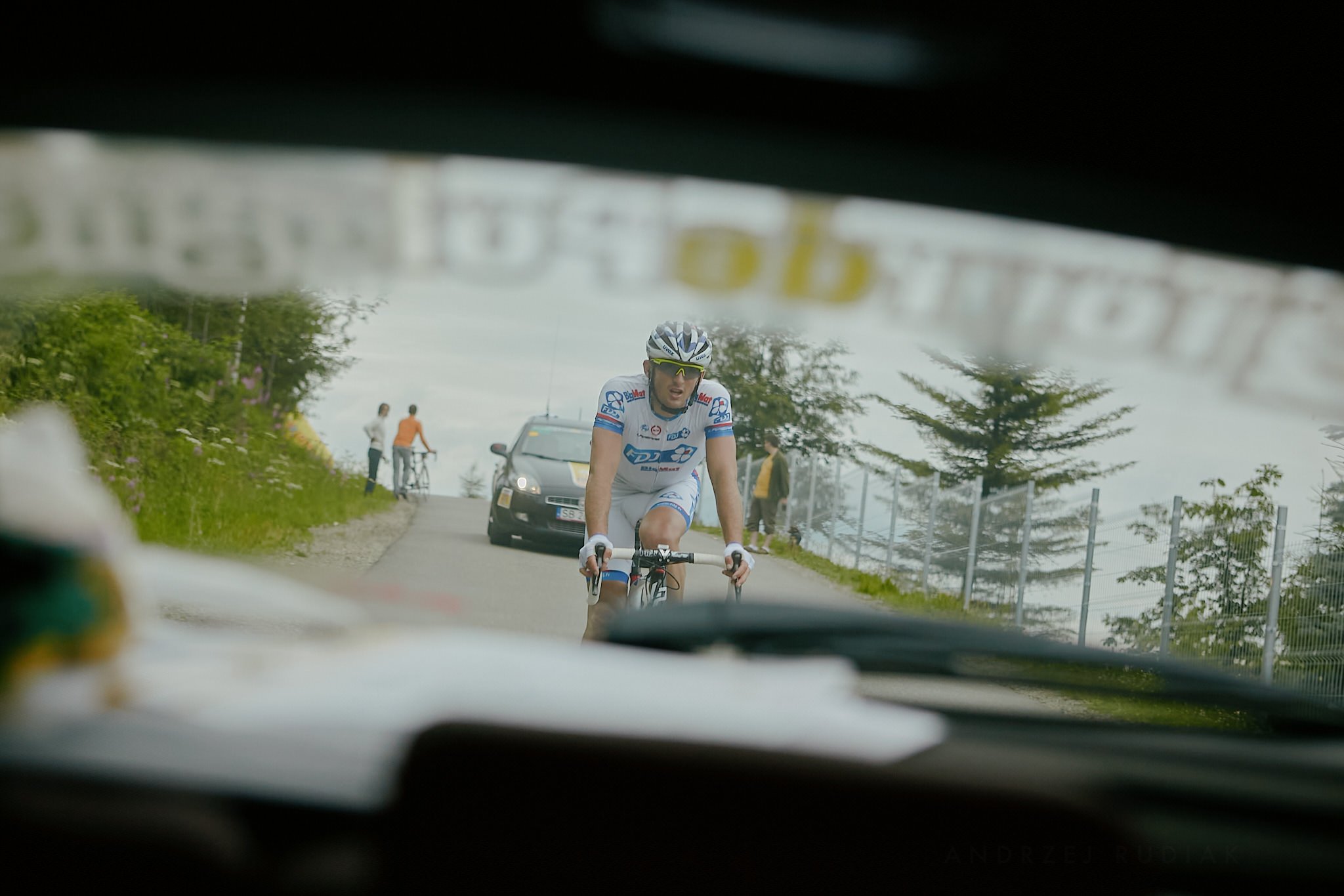 A cyclist in a white and blue racing outfit and helmet riding a bike on a road, with supporters, trees, and cars in the background, viewed from inside a vehicle.