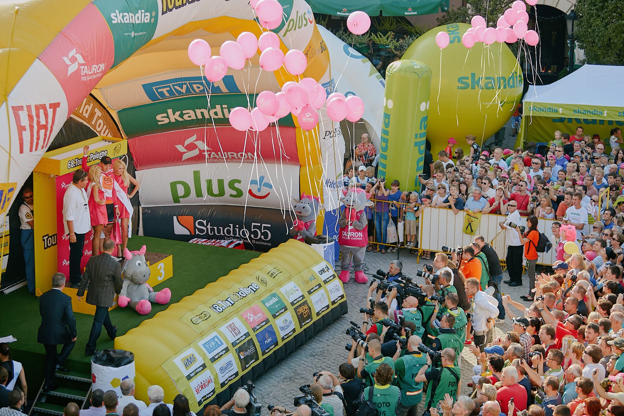 Crowd gathered at an outdoor cycling race event with large inflatable arches and pink balloons, some people dressed in costumes and official jerseys, with photographers capturing the event.