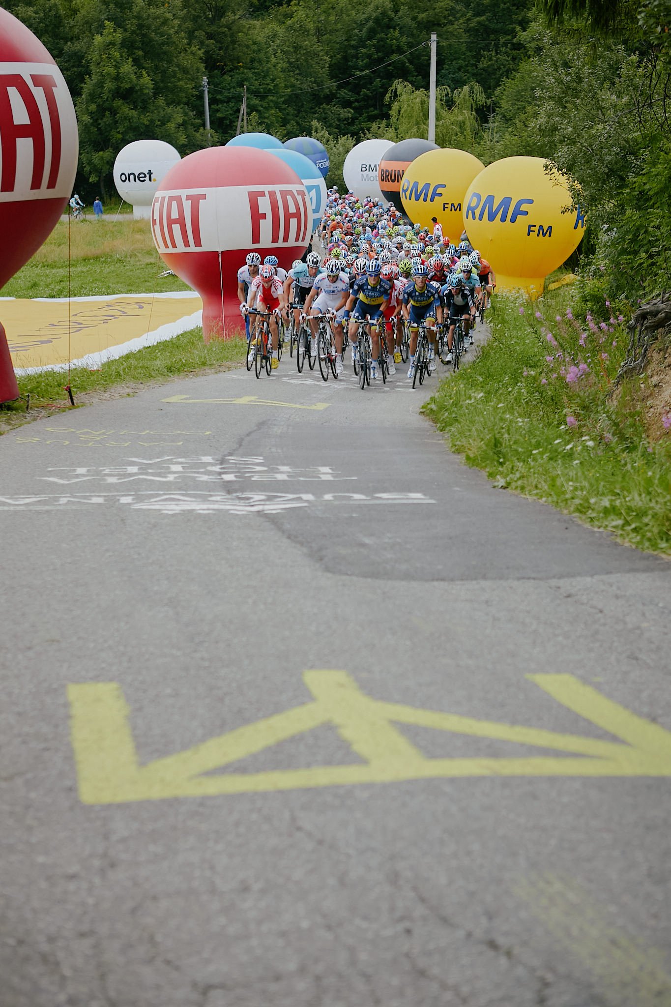 Cyclists participating in a race or event, riding on a narrow road surrounded by trees, with large promotional balloons from brands like FIAT, RMF FM, and others lining the sides.