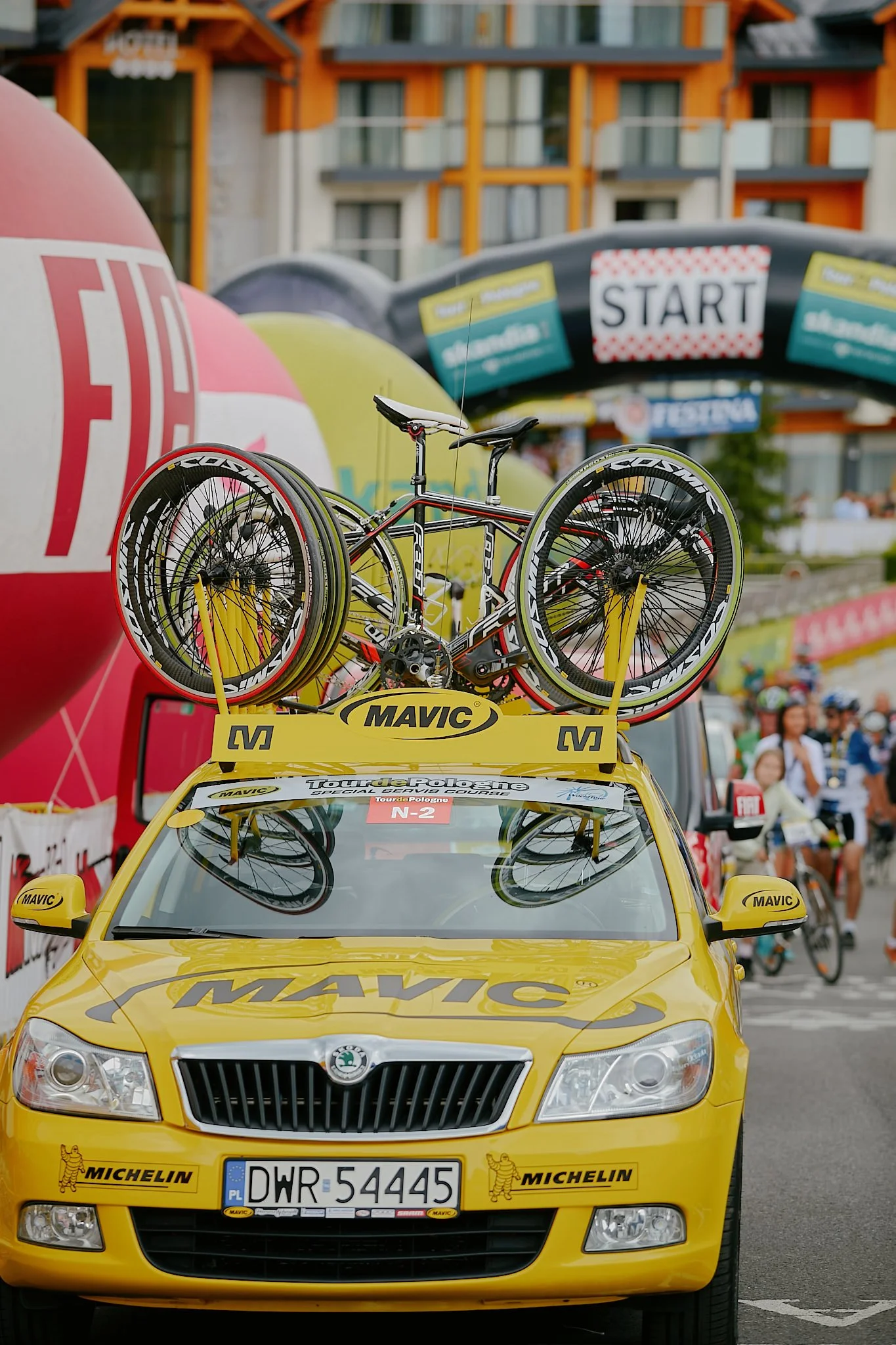 A yellow car with race banners and a bike on the roof rack at a racing event, with people and tents in the background.